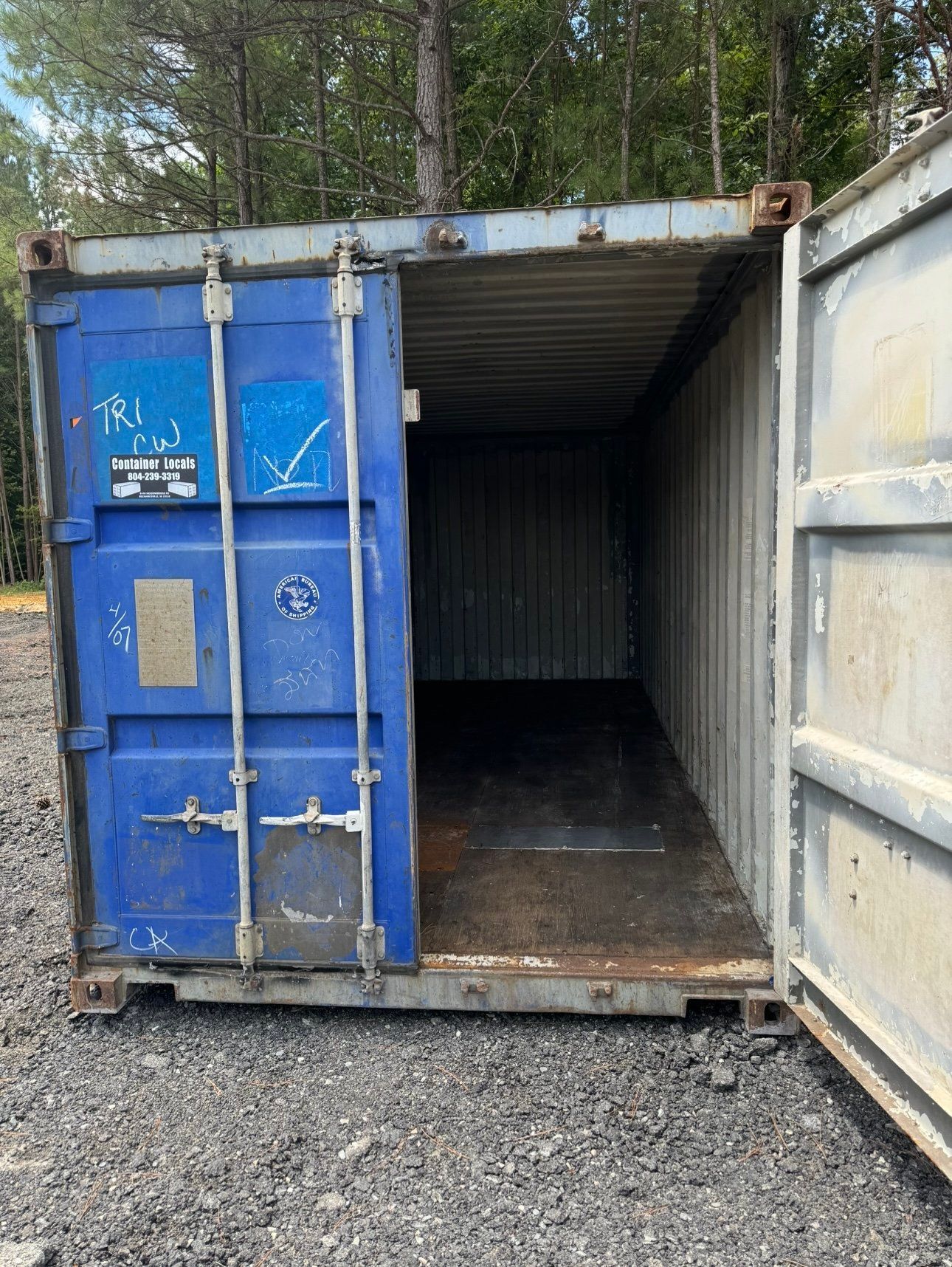 A blue shipping container with the door open is sitting in a gravel lot.