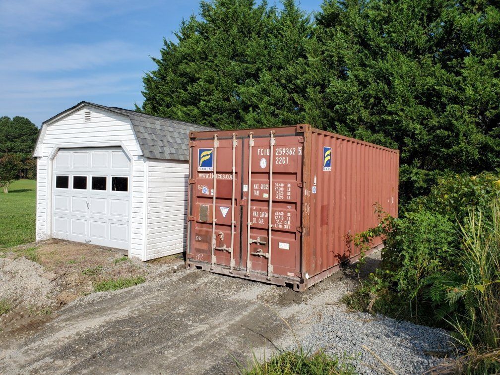 A red shipping container is sitting next to a white garage.