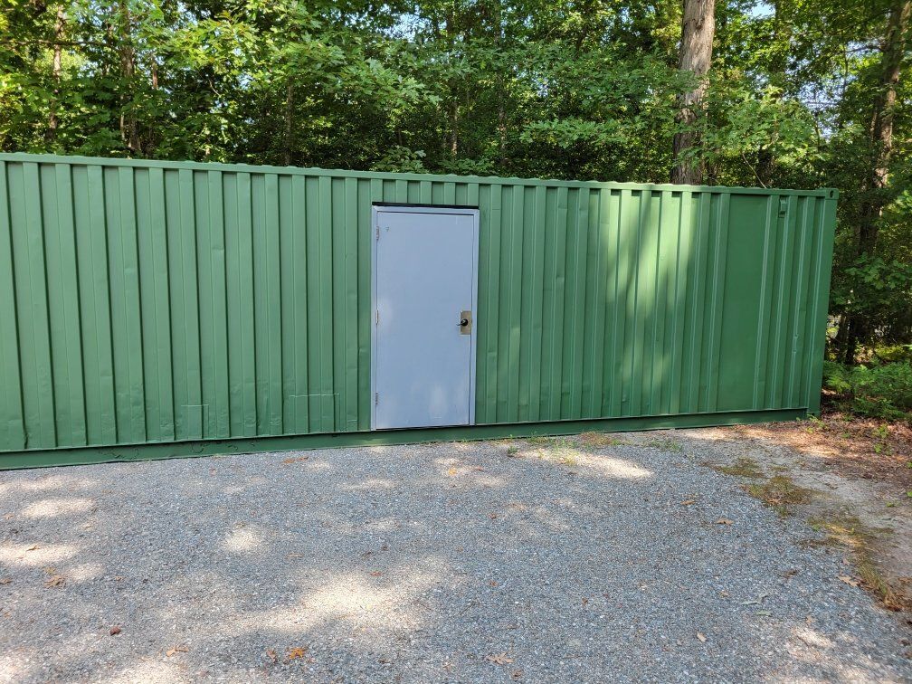 A green building with a white door in the middle of a gravel driveway.