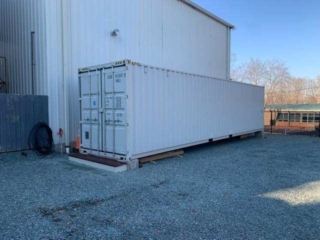 A large white shipping container is parked in a gravel lot next to a building.
