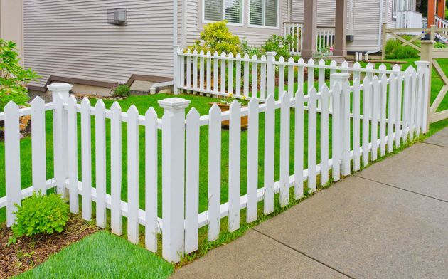 White picket fence enclosing a small green yard beside a sidewalk and house exterior