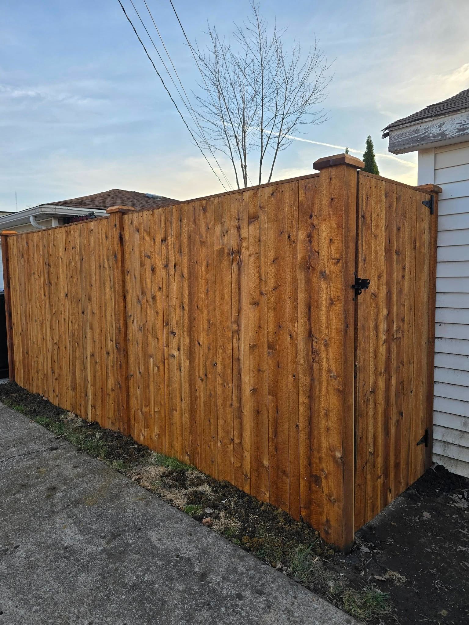 Wooden privacy fence with gate beside a house, on a gravel driveway under a clear sky