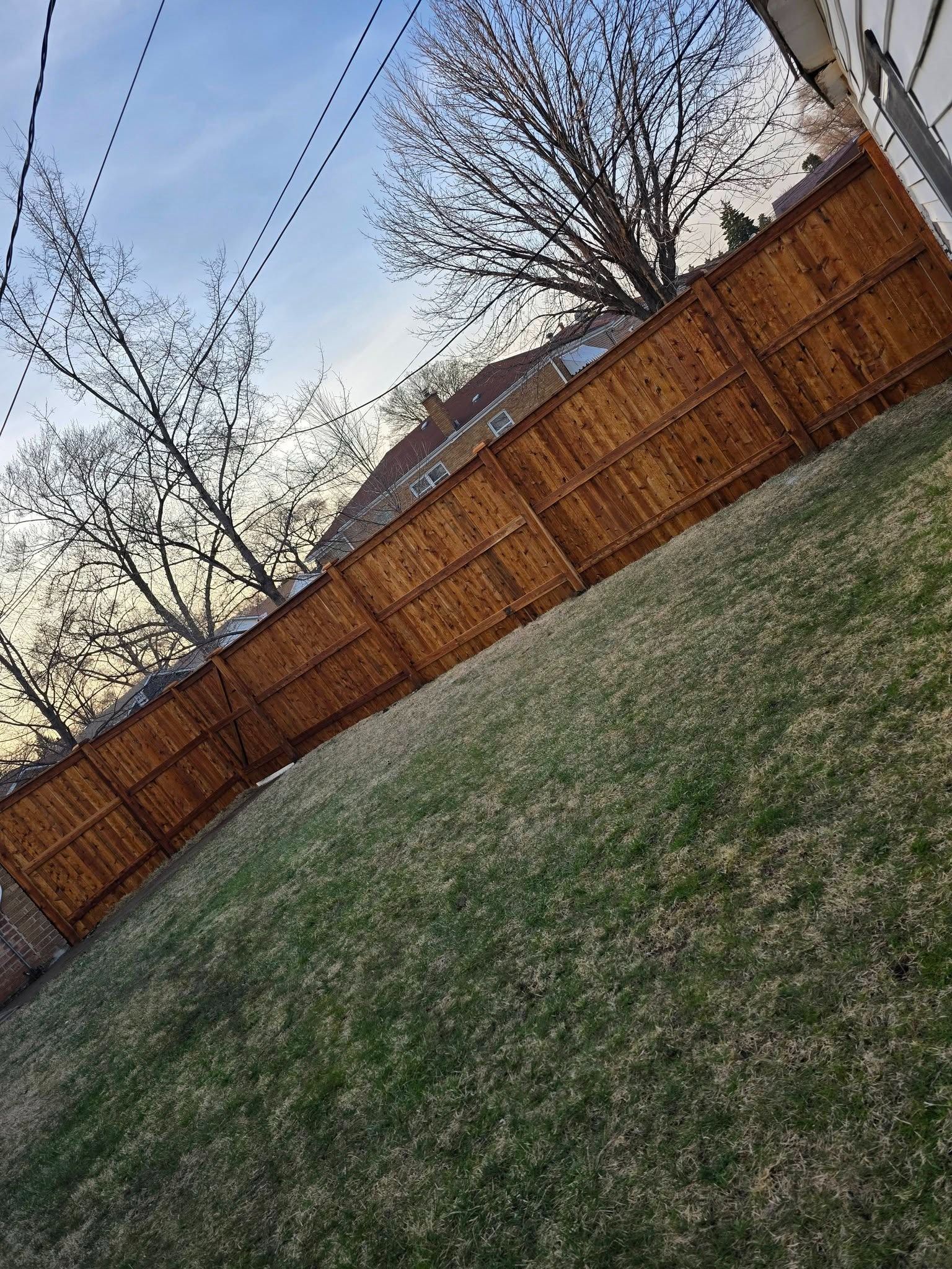 Tilted view of a grassy backyard with a wooden fence, leafless trees, and a house roof in the background