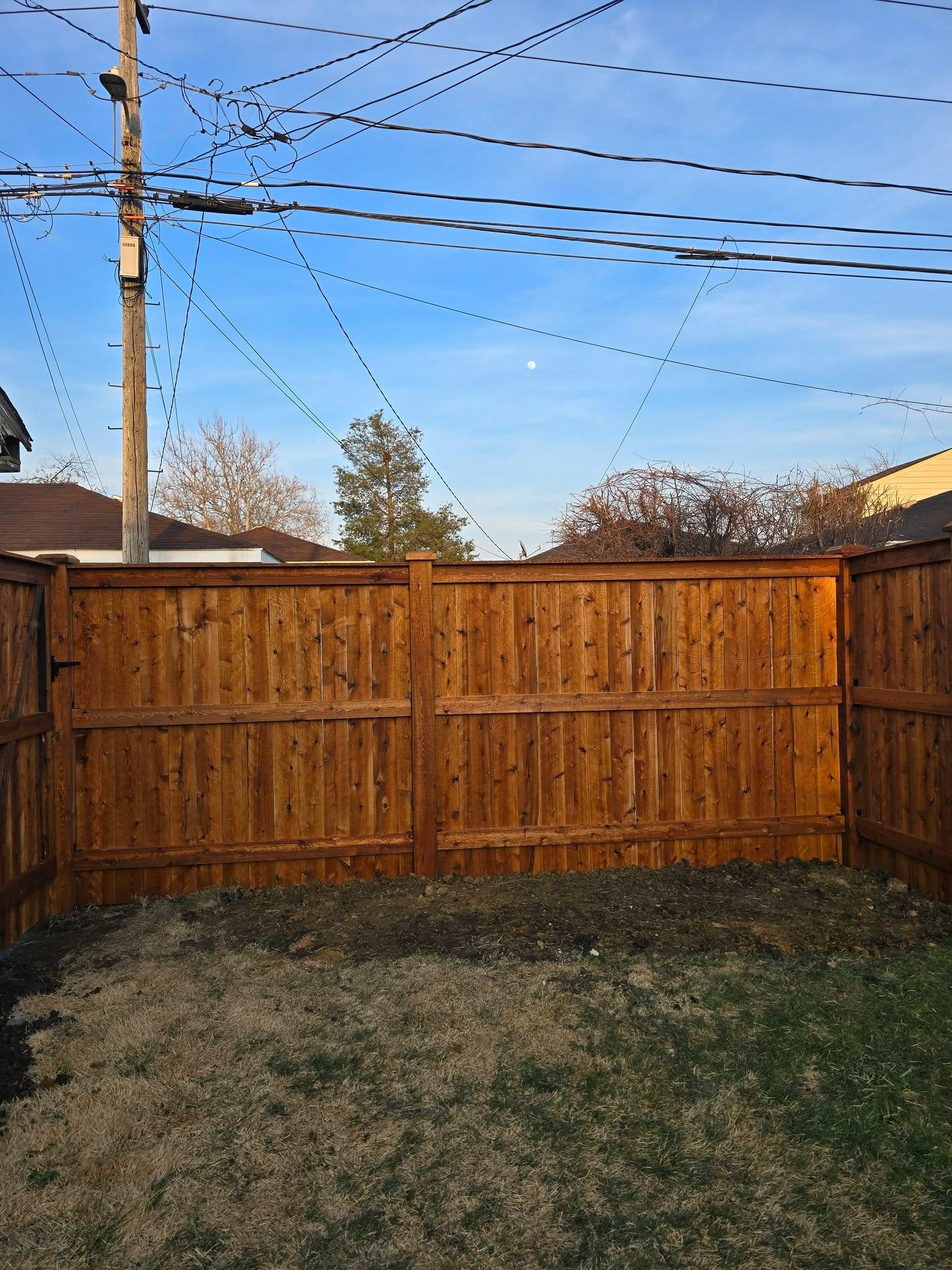 Backyard with a wooden fence, patchy grass, utility poles, and overhead power lines at dusk