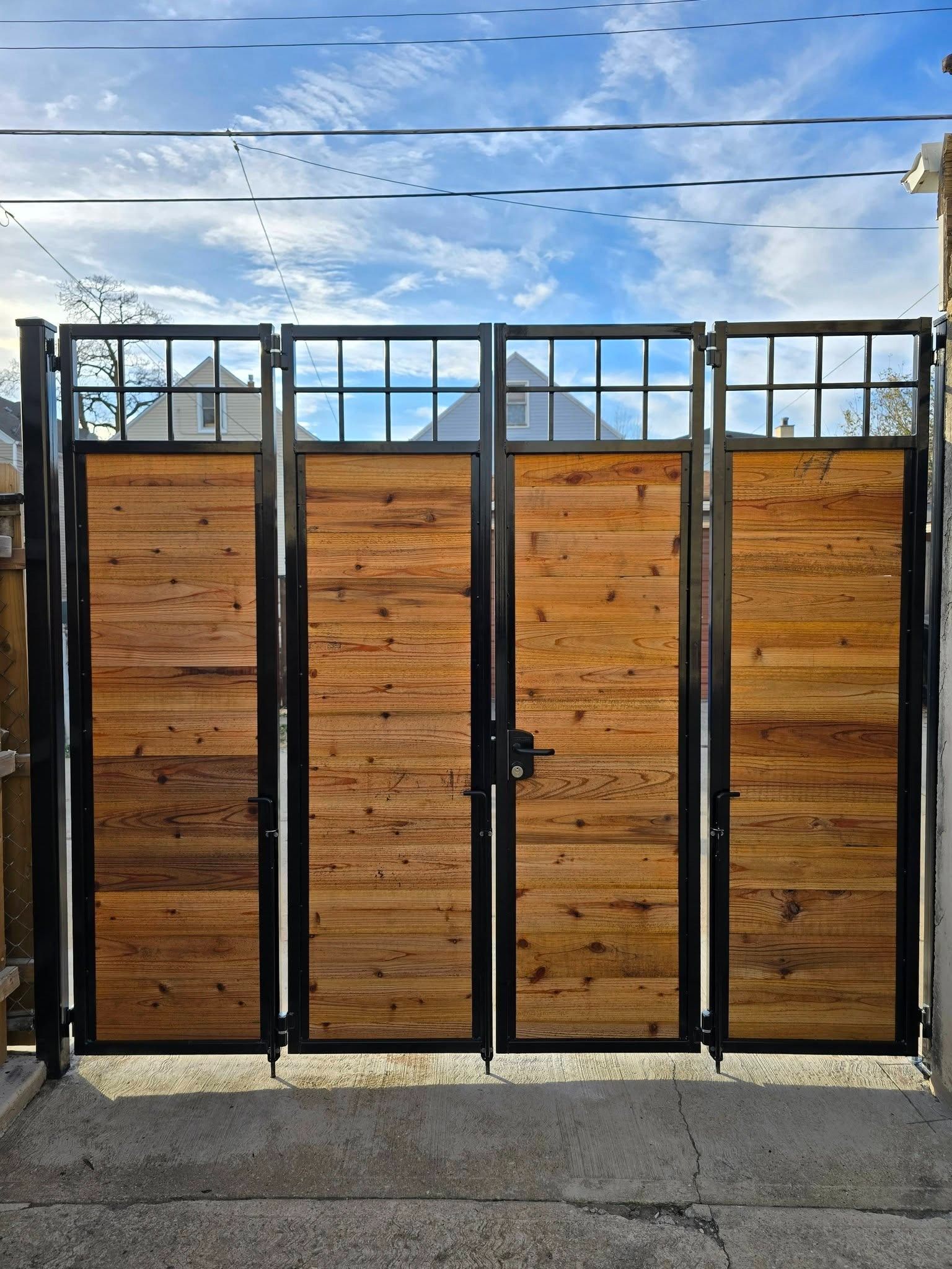 Black metal-and-wood gate with four panels under a cloudy sky, viewed from the street