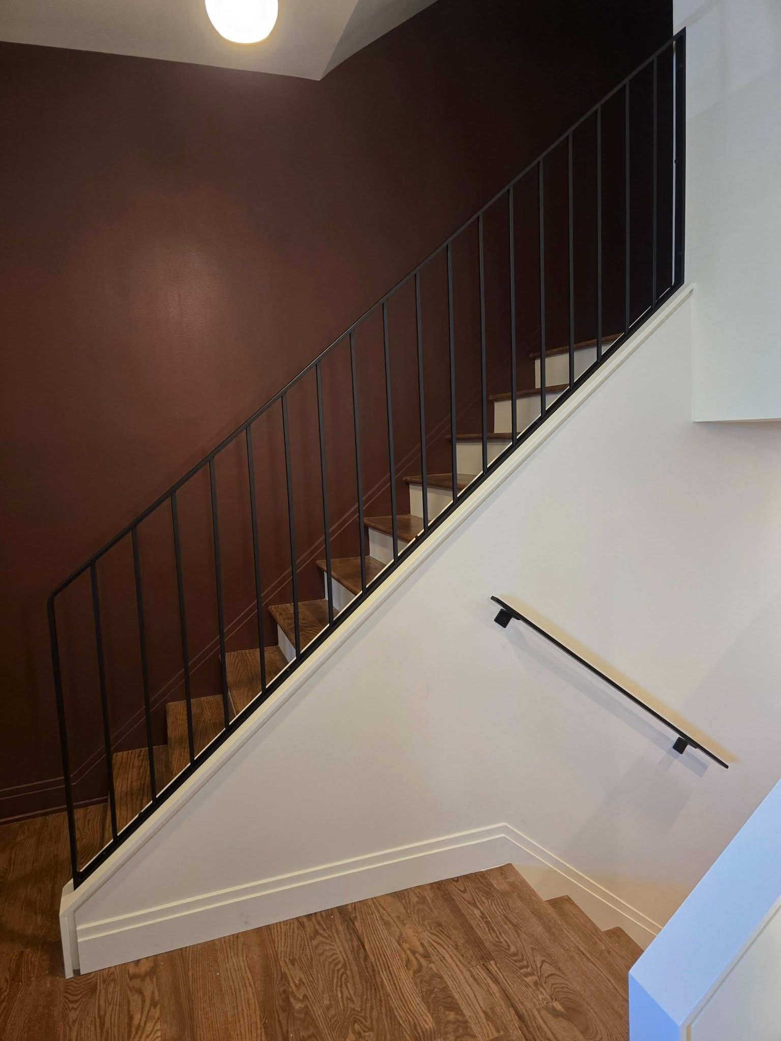 Interior staircase with dark railing, brown wall, white trim, and wood flooring