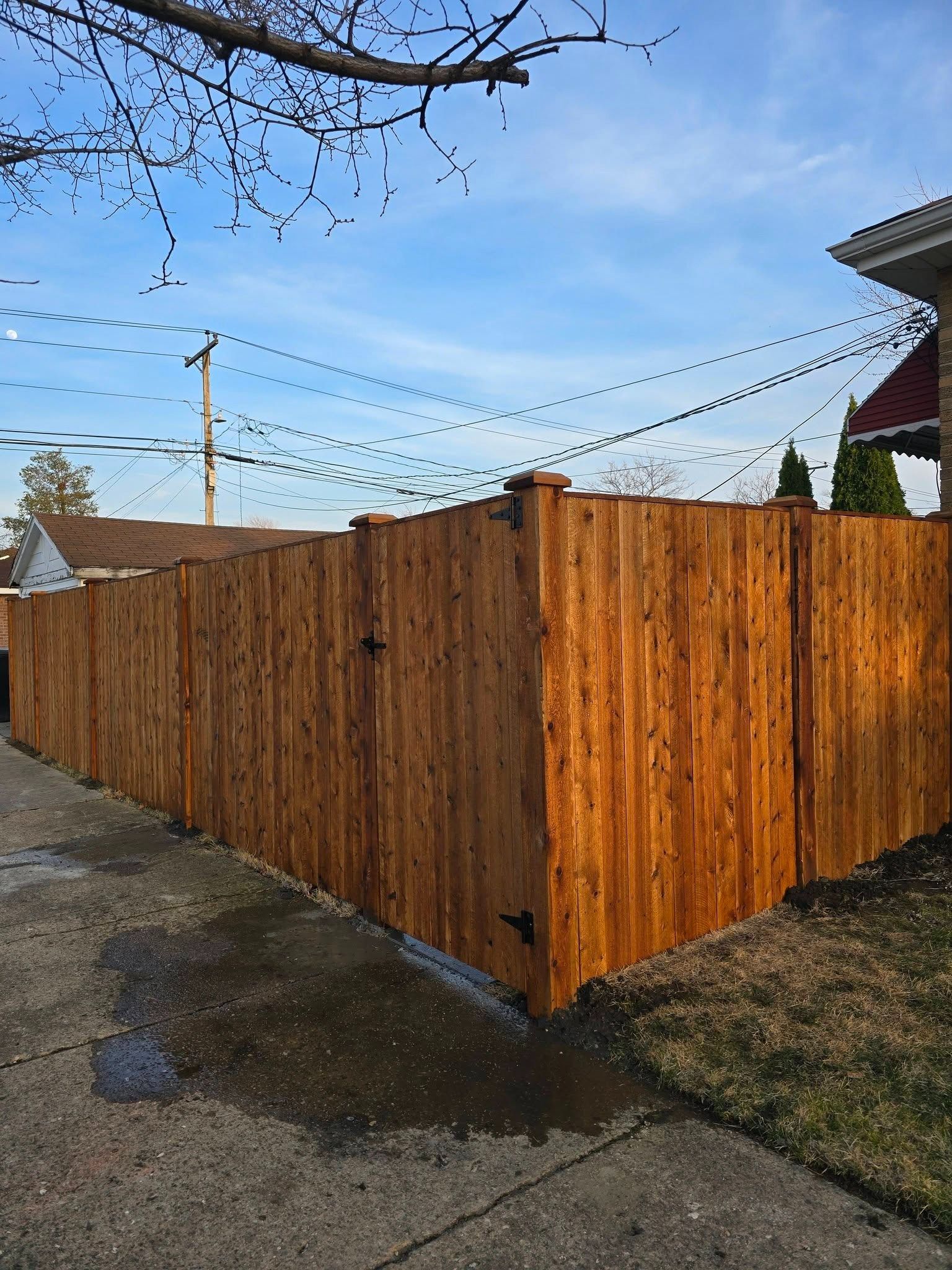 Wooden privacy fence along a wet sidewalk at a street corner under a blue sky.