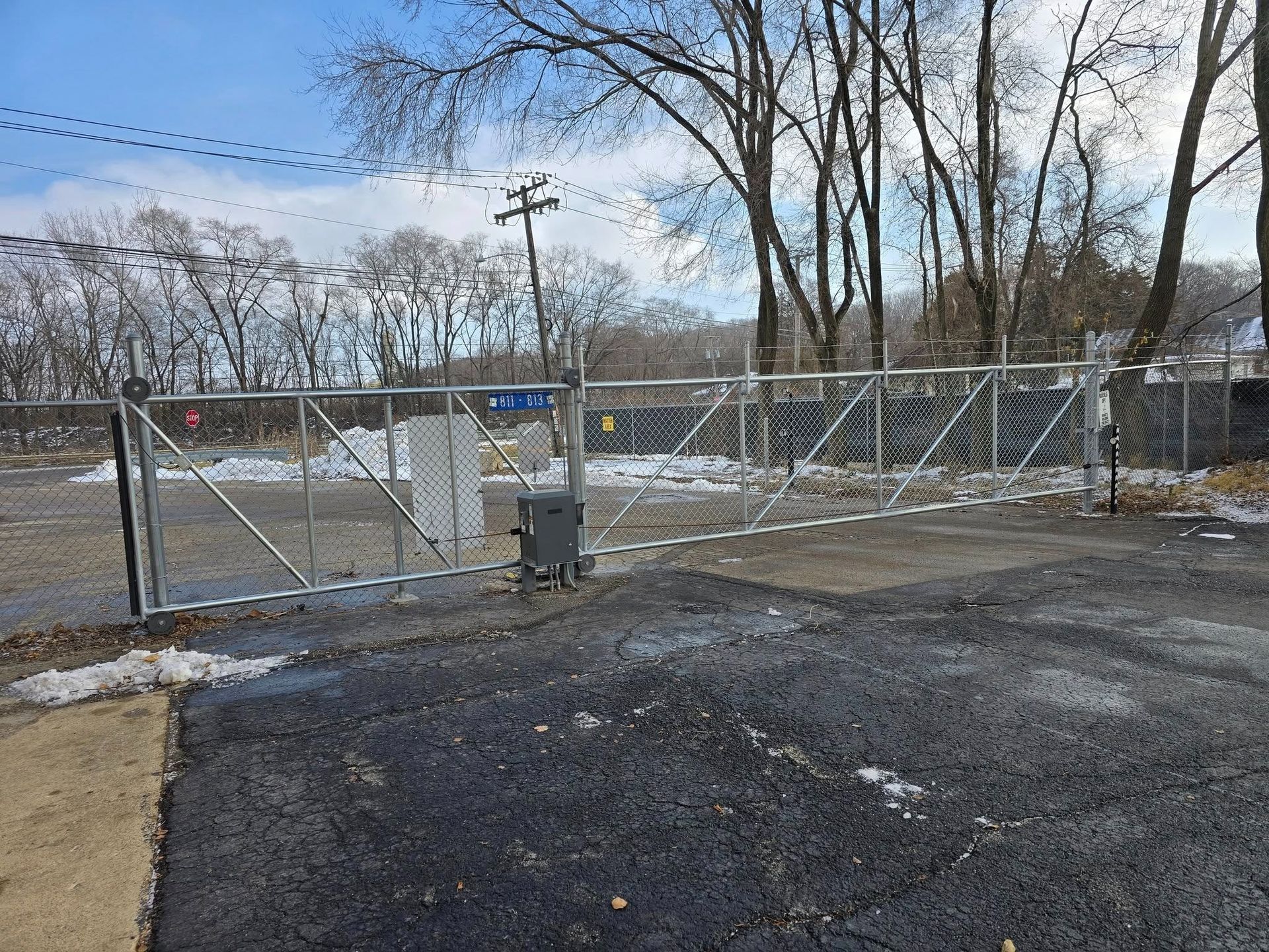 Closed chain-link gate across a snowy driveway with utility poles and bare trees behind it