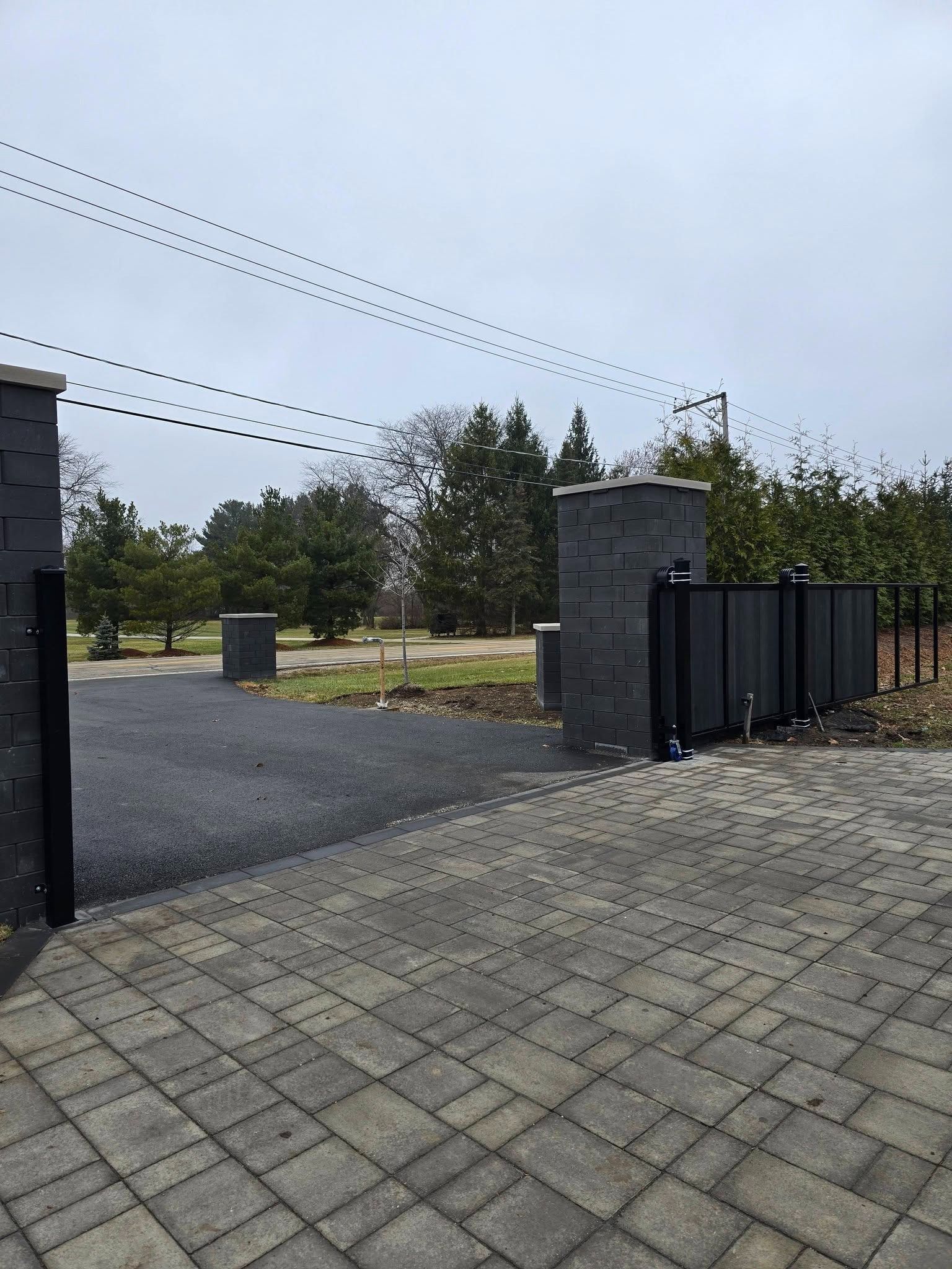 Gravel driveway with stone gate pillars, black fence, and trees under a cloudy sky