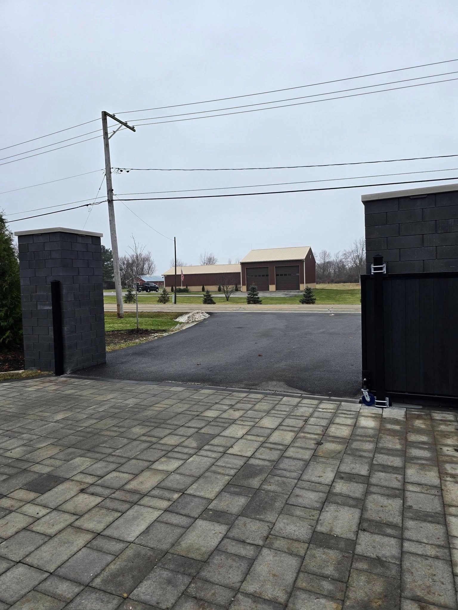 Paved driveway entrance between brick pillars, opening to a road and a house in the distance.