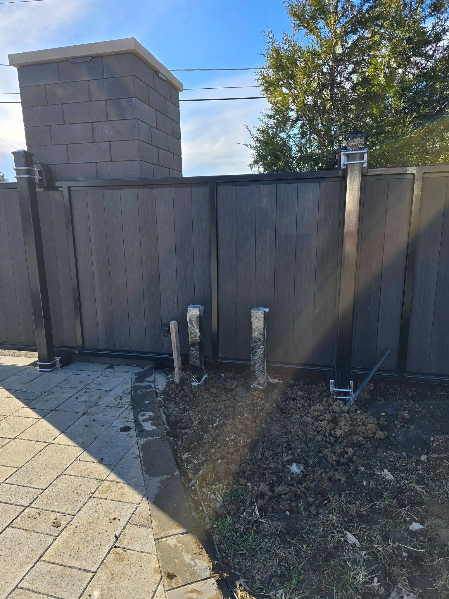Wooden fence and gate beside a stone pillar, with a paved patio and sunny yard.
