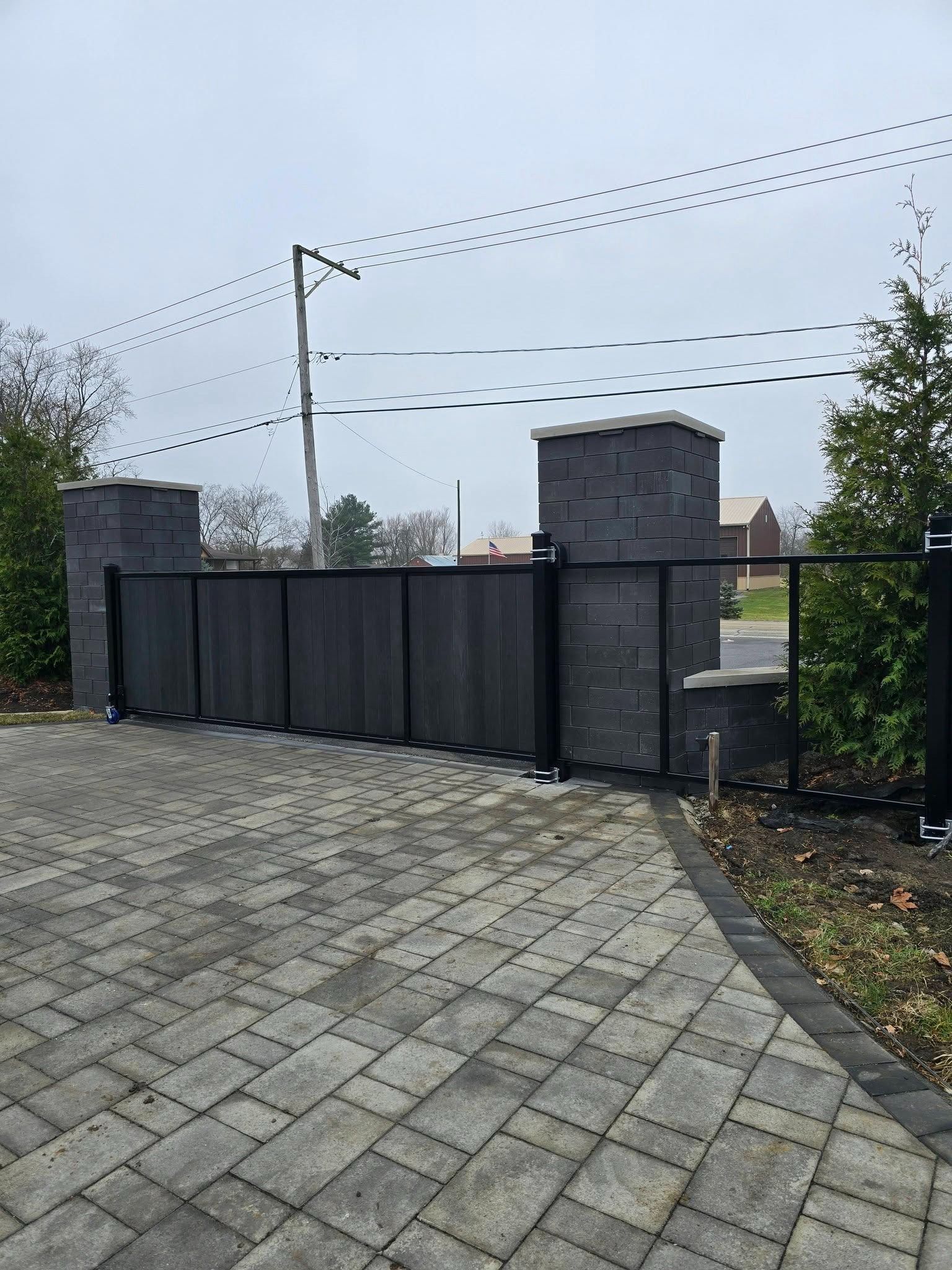 Black metal gate between stone pillars at the end of a paved driveway under a cloudy sky