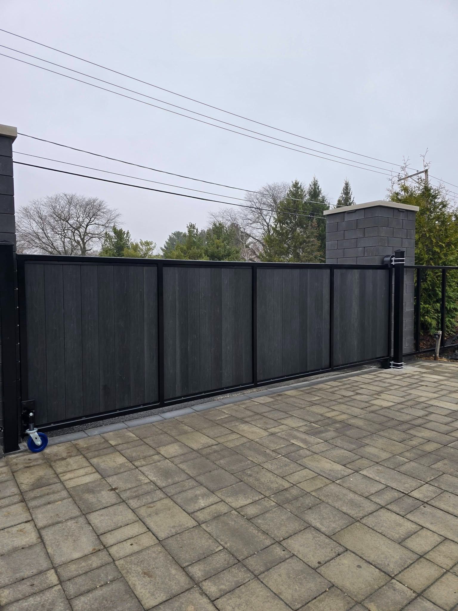 Dark sliding driveway gate beside a paved courtyard and brick pillar under overcast sky