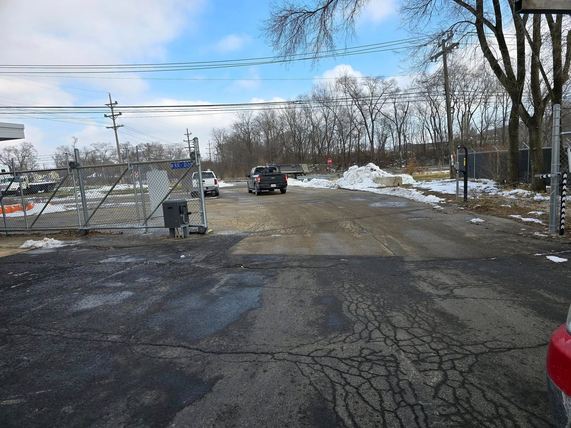 Snowy cracked road leading to a closed gate with a black SUV in the distance and bare trees