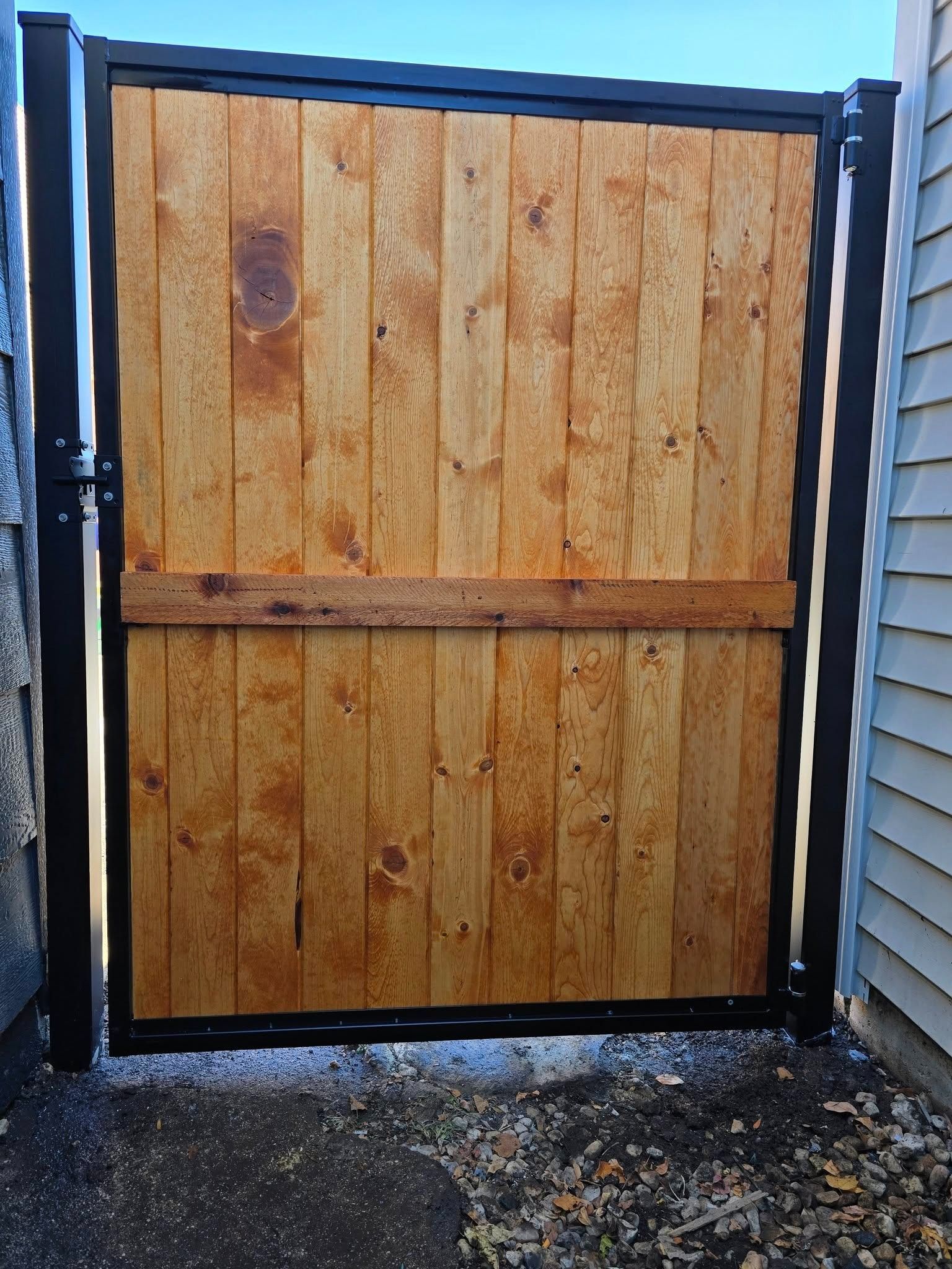 Wooden gate with black metal frame between stone walls, viewed from outside on a gravel surface.