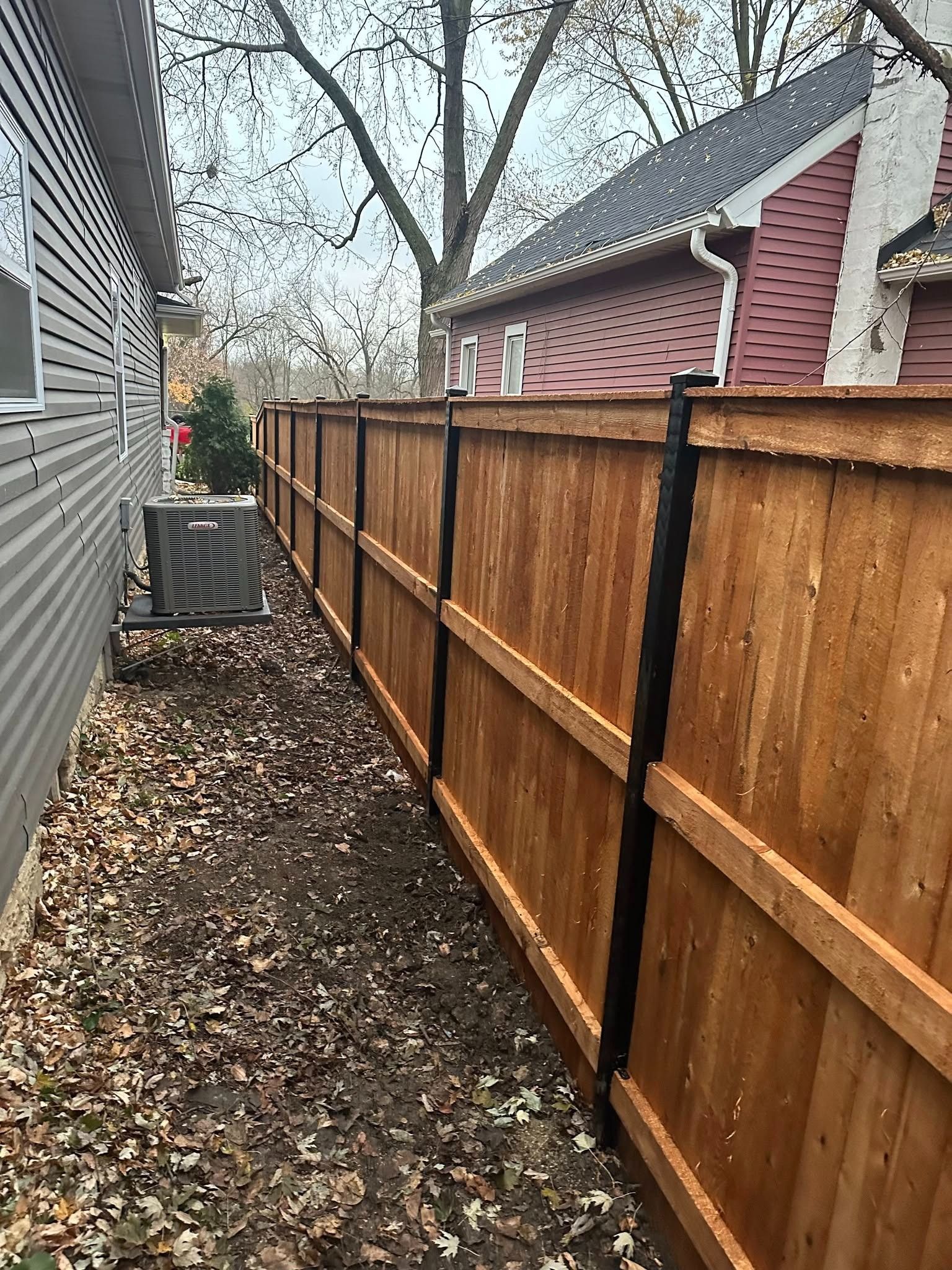 Narrow side yard with brown wooden fence, gray siding, fallen leaves, and houses under bare trees
