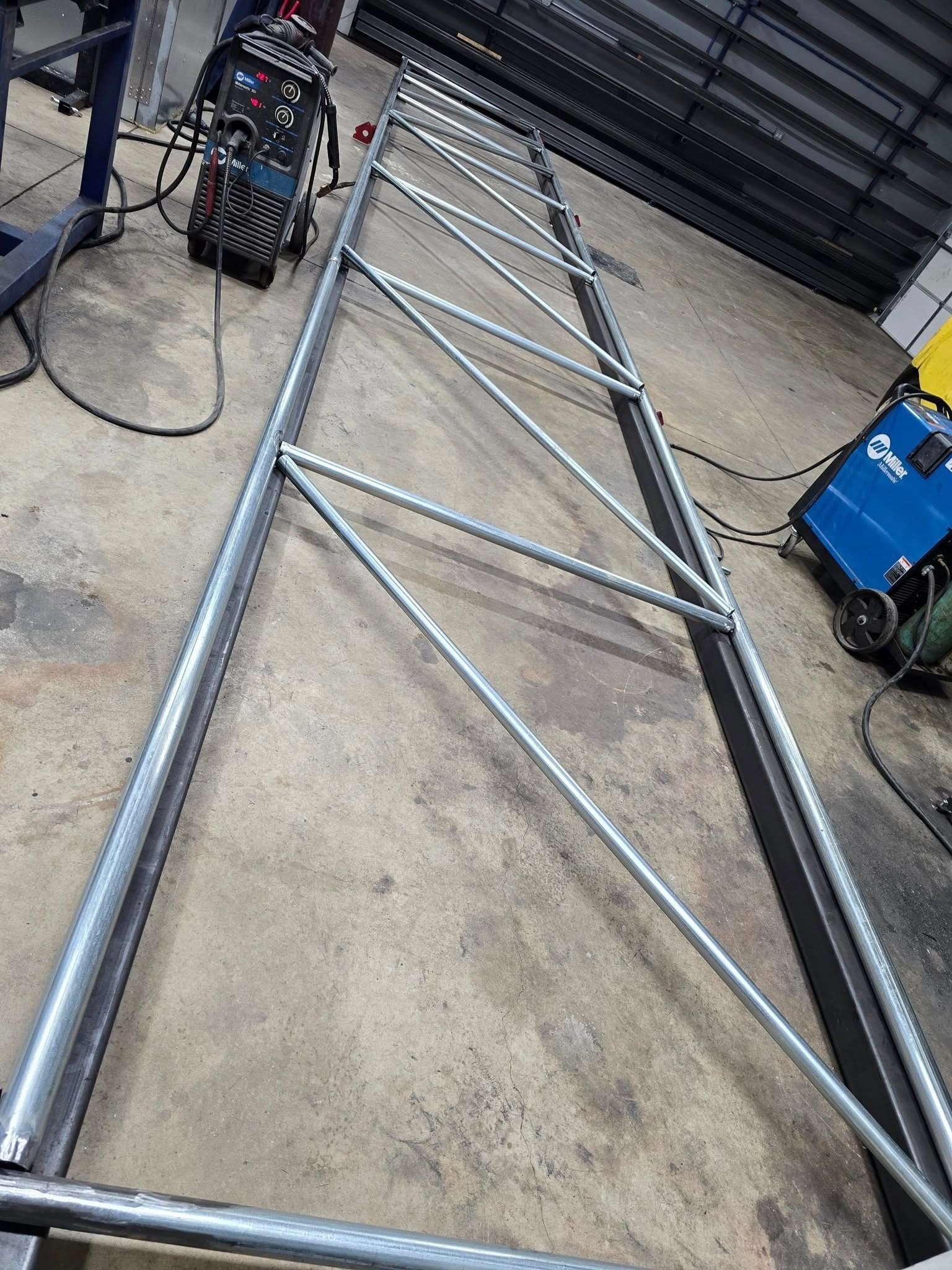Metal truss frame on a concrete floor, with welding equipment and a blue machine nearby in a workshop.