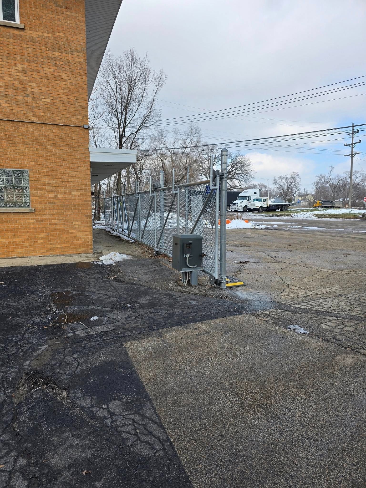 Brick building edge beside a chain-link fence, gravel lot, and cloudy winter sky.
