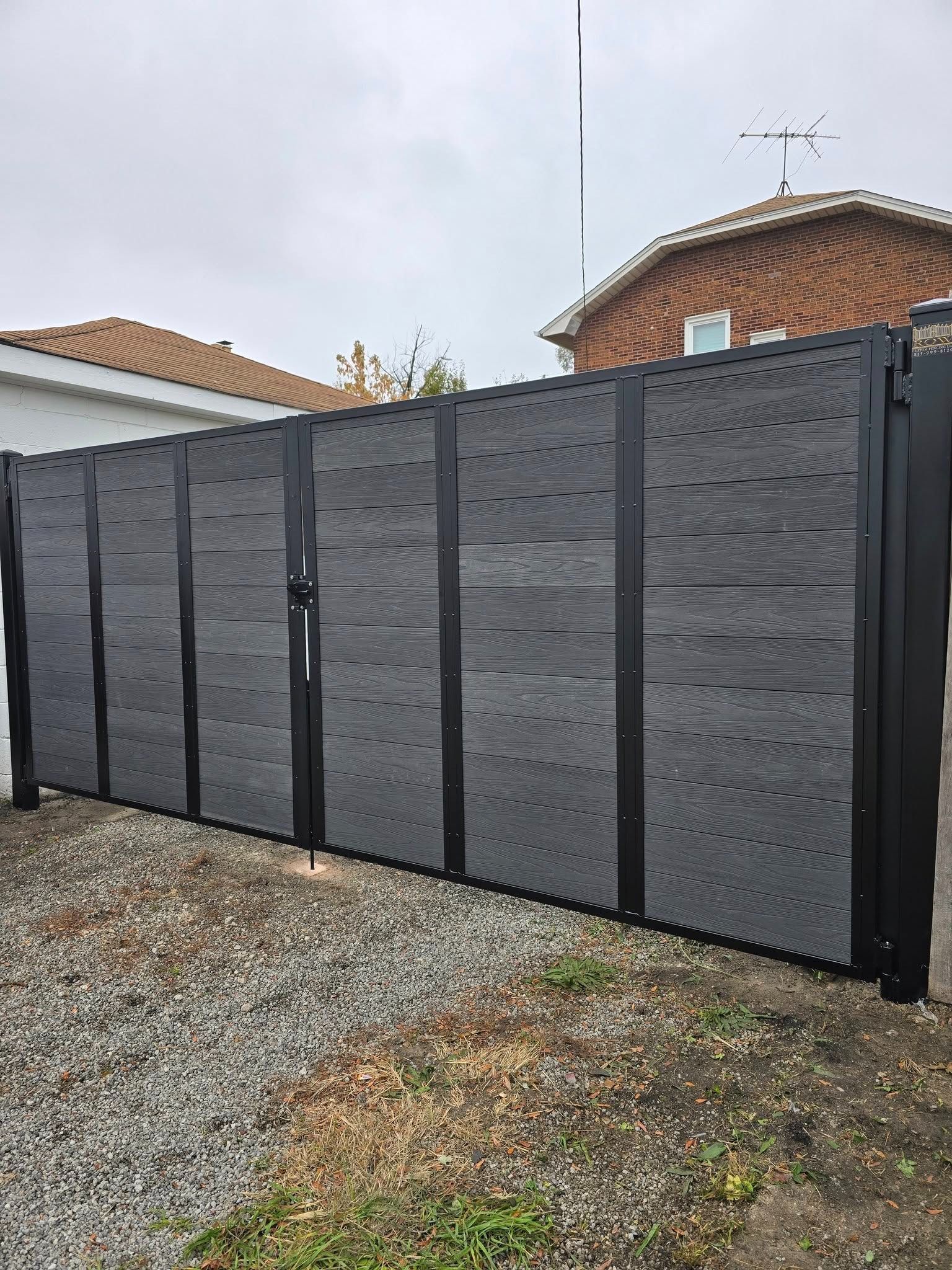 Tall dark gray fence panels beside a gravel driveway in front of a brick house.