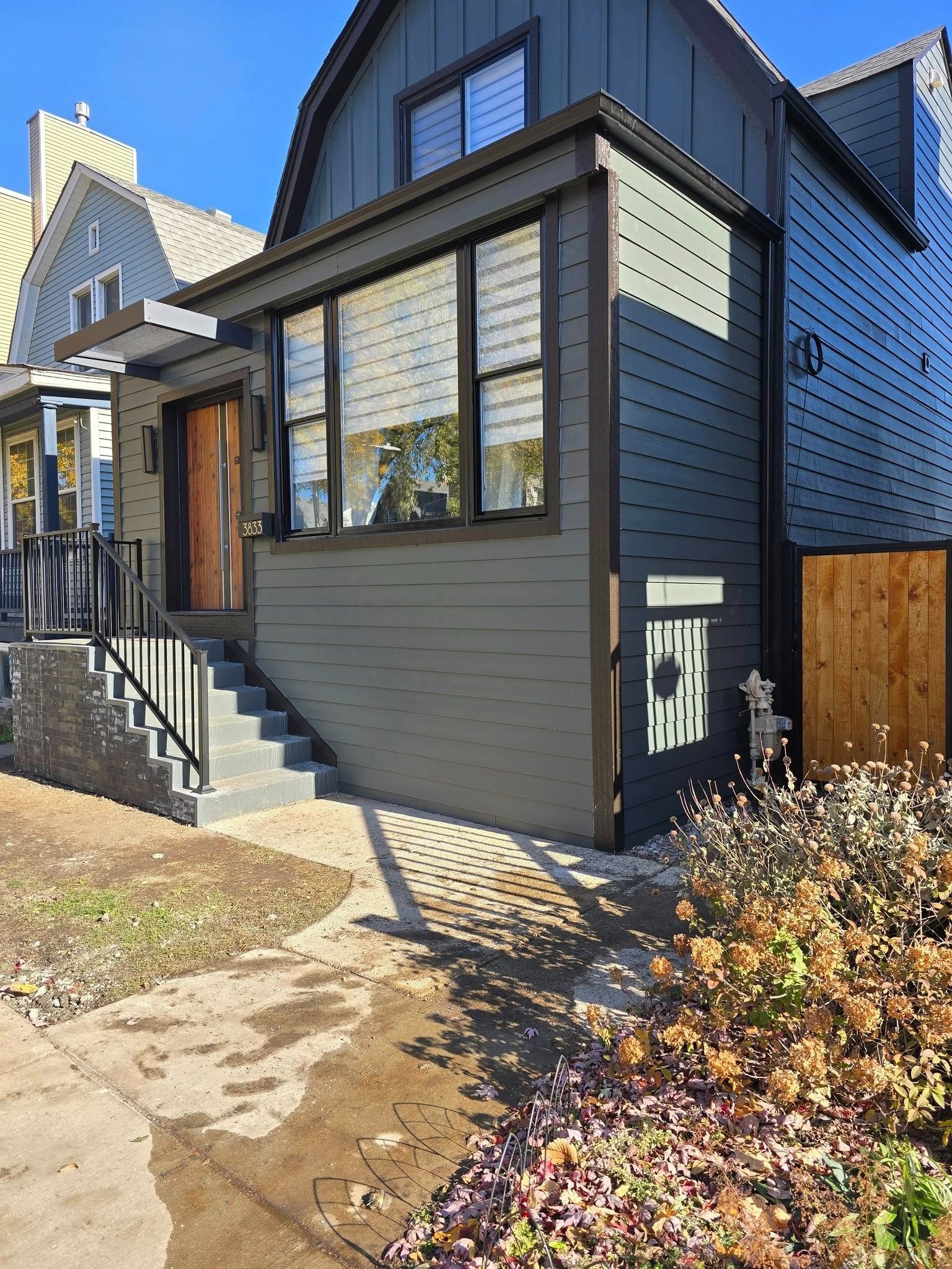 Modern gray house exterior with front steps, black trim, and small garden bed in sunlight