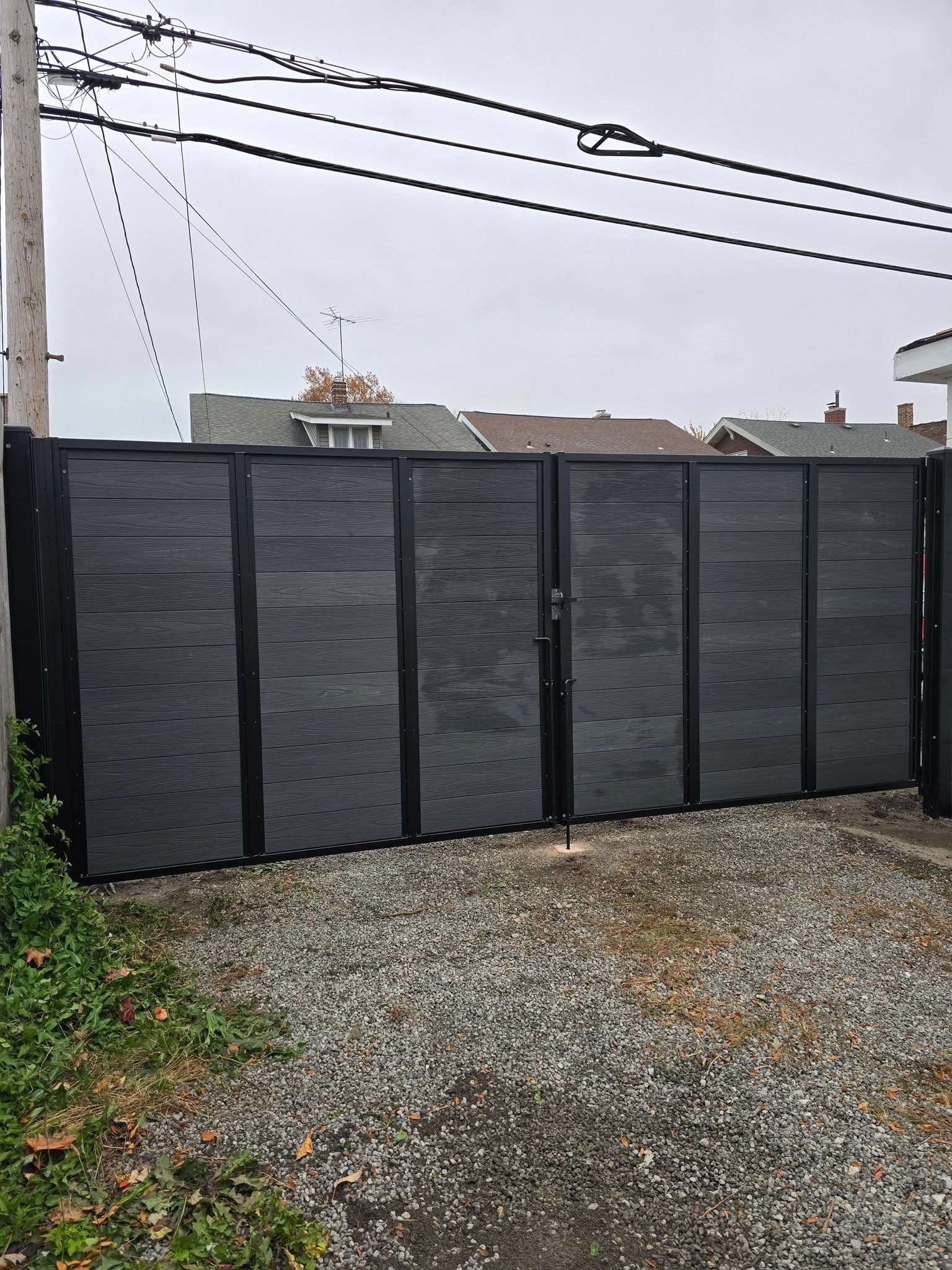 Dark gray fence panels along a gravel driveway beside utility poles and overhead wires under a cloudy sky