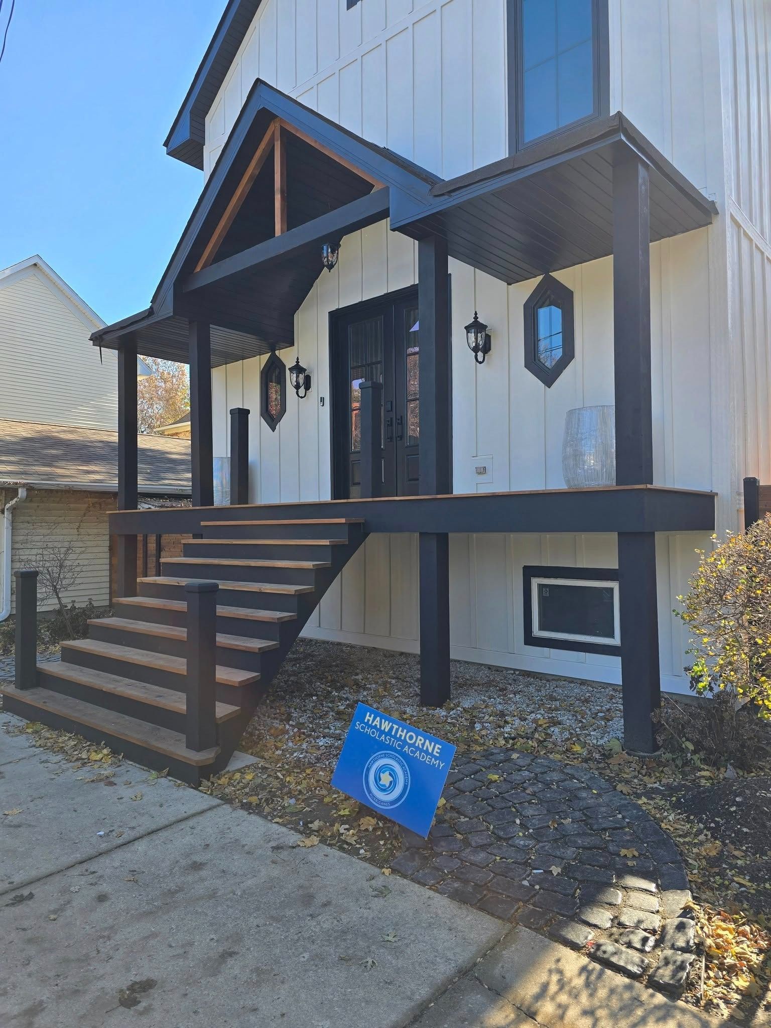 White-and-black building entrance with wooden steps, porch lights, and a blue sign leaning in front