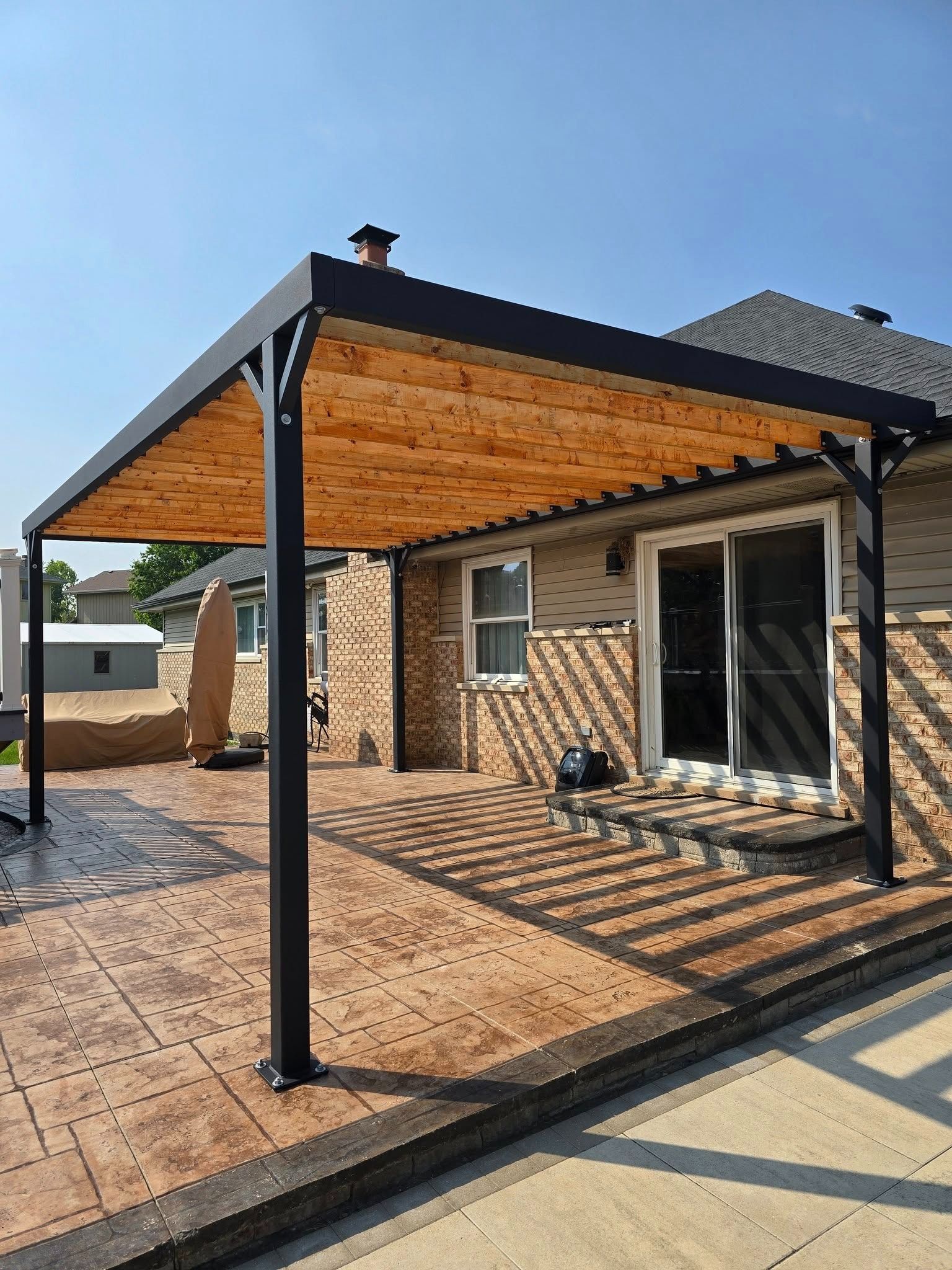 Covered patio with black metal posts and wood ceiling beside a brick house, in bright sunlight.