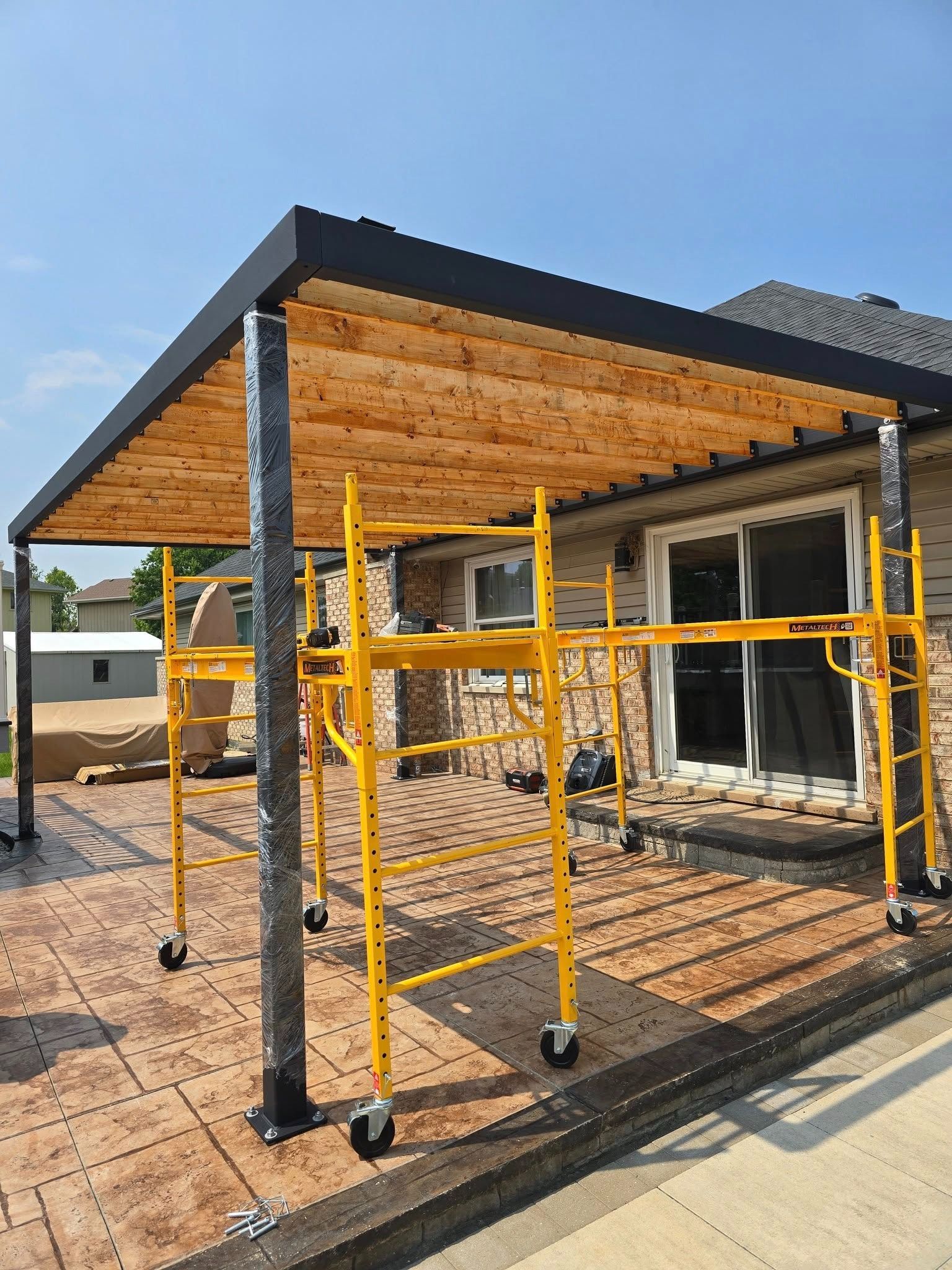 Partially built porch with yellow scaffolding and a black roof beside a brick walkway