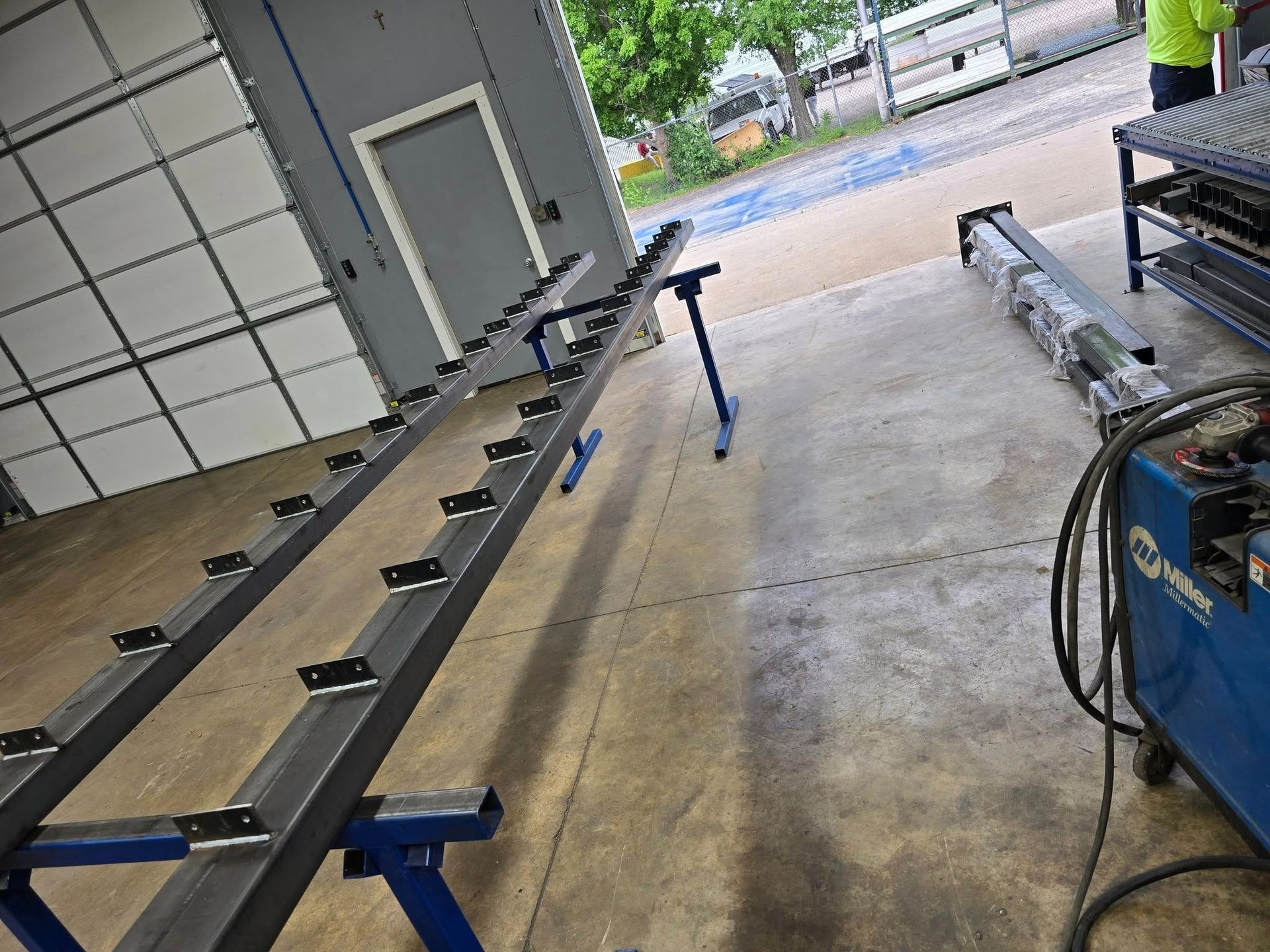 Metal worktable frame in a workshop, with clamps, blue supports, and a welding machine nearby.