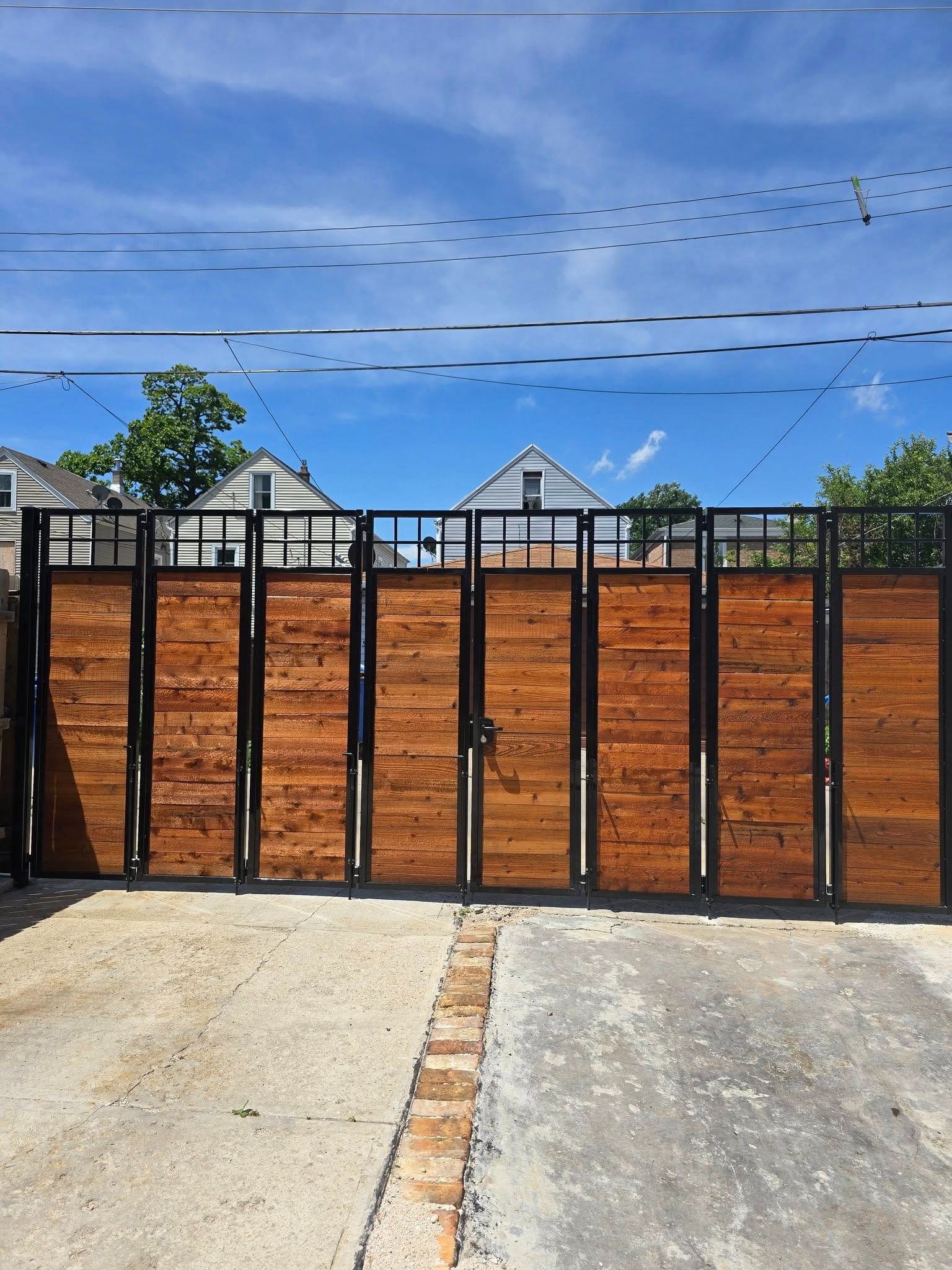 Closed black metal gate in front of a wooden fence and house, with driveway and blue sky overhead