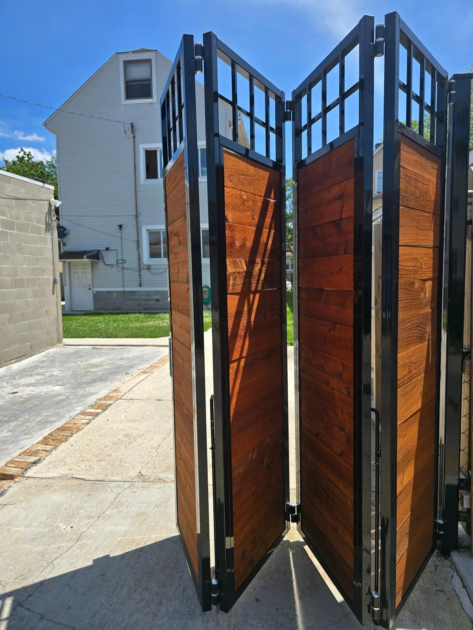 Black metal and wood folding gate standing open on a sidewalk near a house, with a driveway and lawn behind.