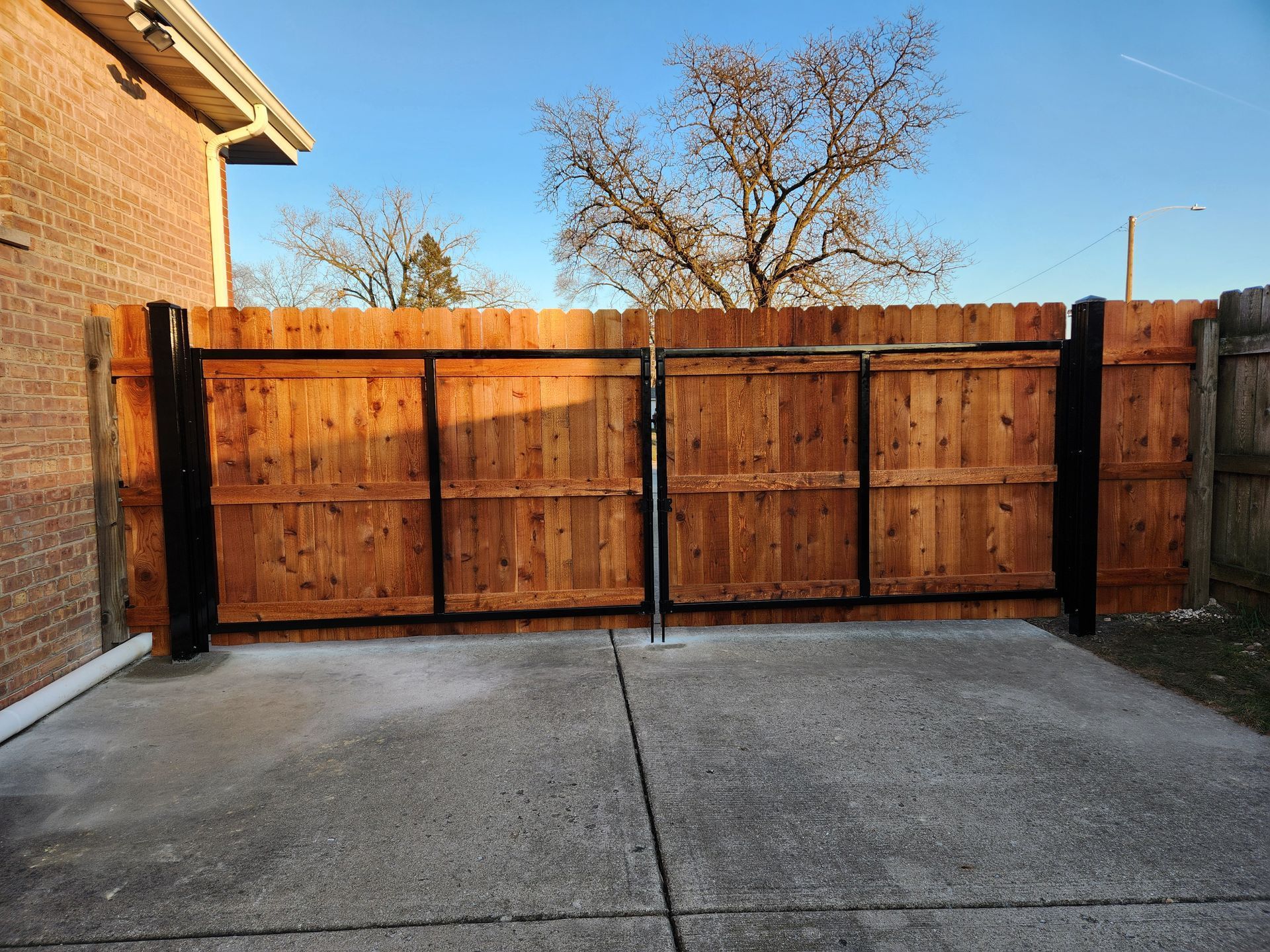 Wooden backyard fence with black posts beside a concrete driveway and brick house at sunset