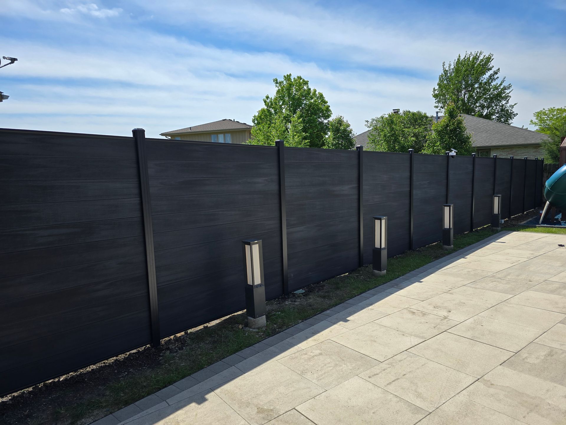 Black wooden fence along a sidewalk under a blue sky, with trees and rooftops in the background
