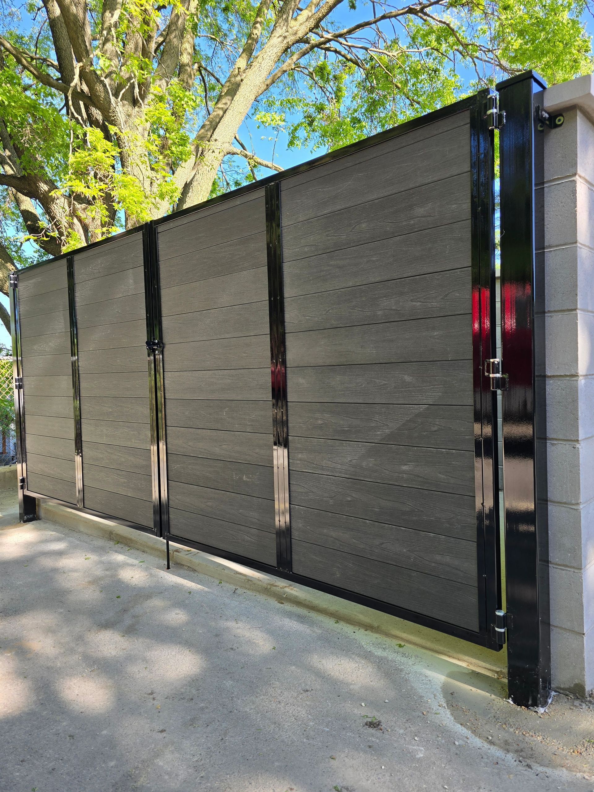 Black metal gate panels beside a paved path under leafy trees in bright daylight
