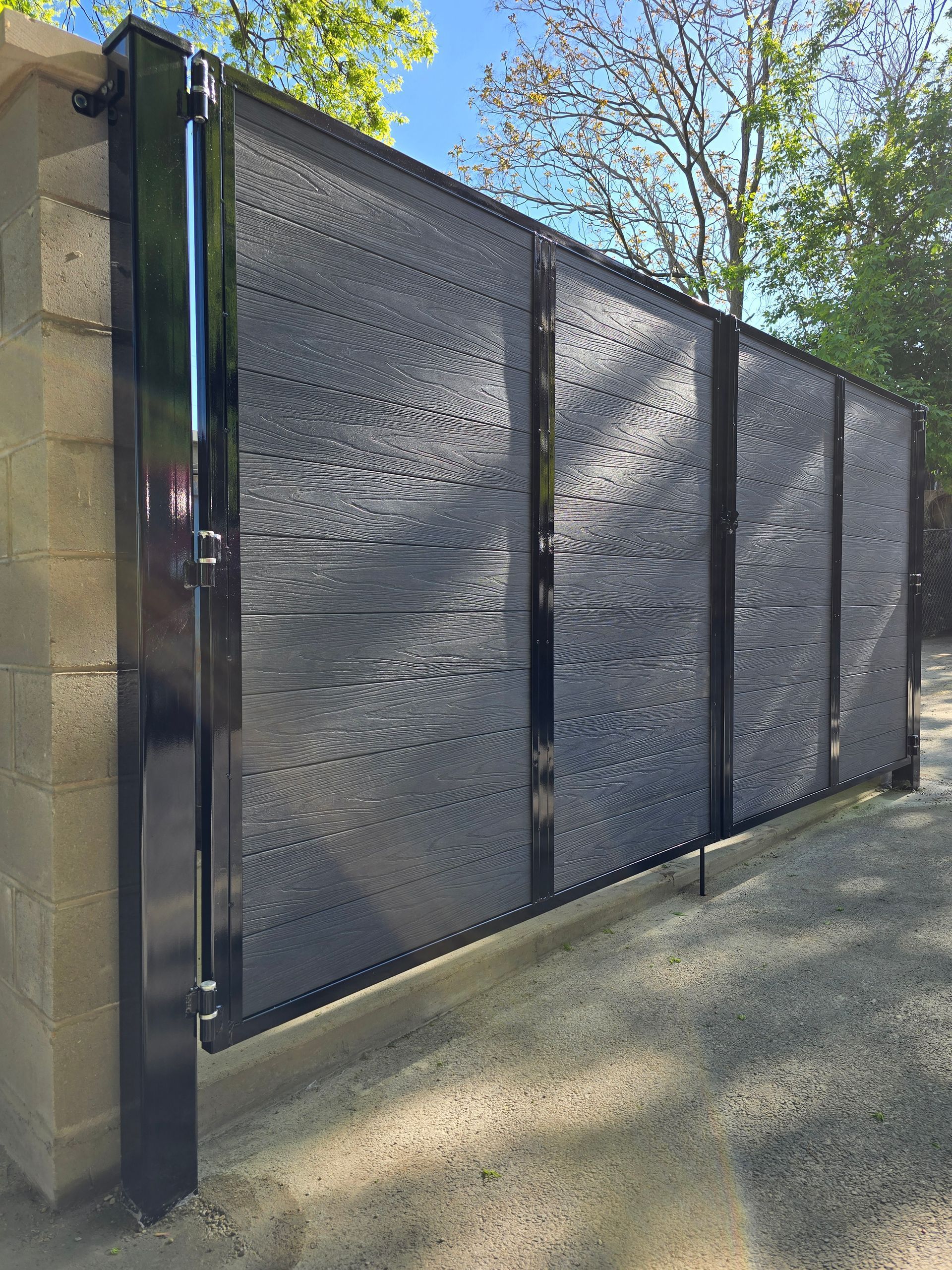 Black metal mesh gate along a stone wall in a sunny outdoor driveway