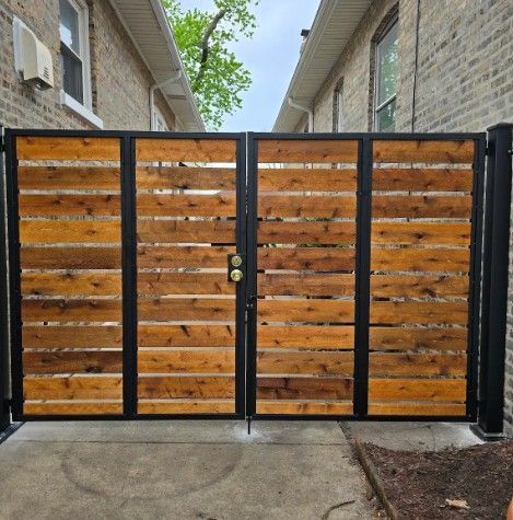 Black metal gate with horizontal wooden slats between two brick buildings and a concrete path.