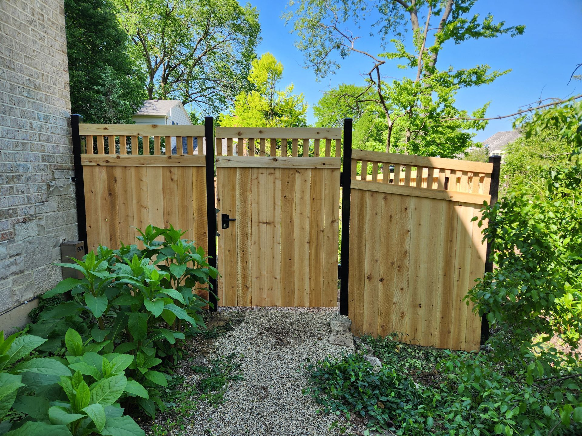 Wooden garden gate and fence beside a stone wall, with gravel path and green shrubs under a bright blue sky