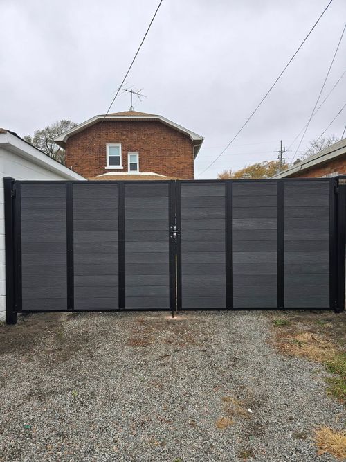 Dark gray driveway gate with black posts in front of a brick house and gravel yard