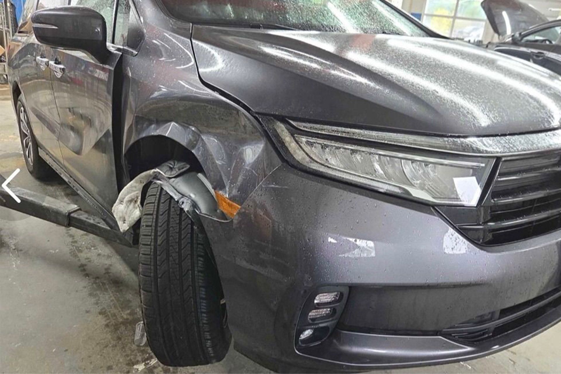 Gray SUV with front-left collision damage, bent wheel, and dented fender in a garage