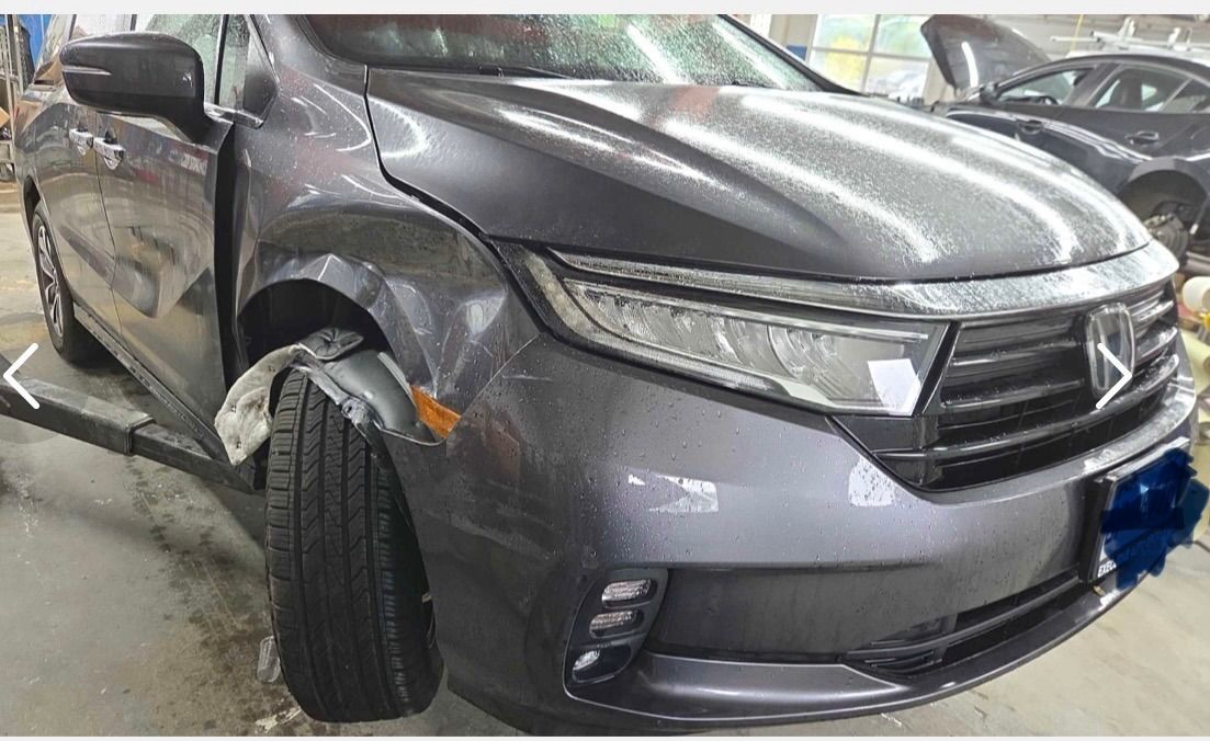 Gray sedan with front-end damage and a bent left wheel in a garage or repair shop