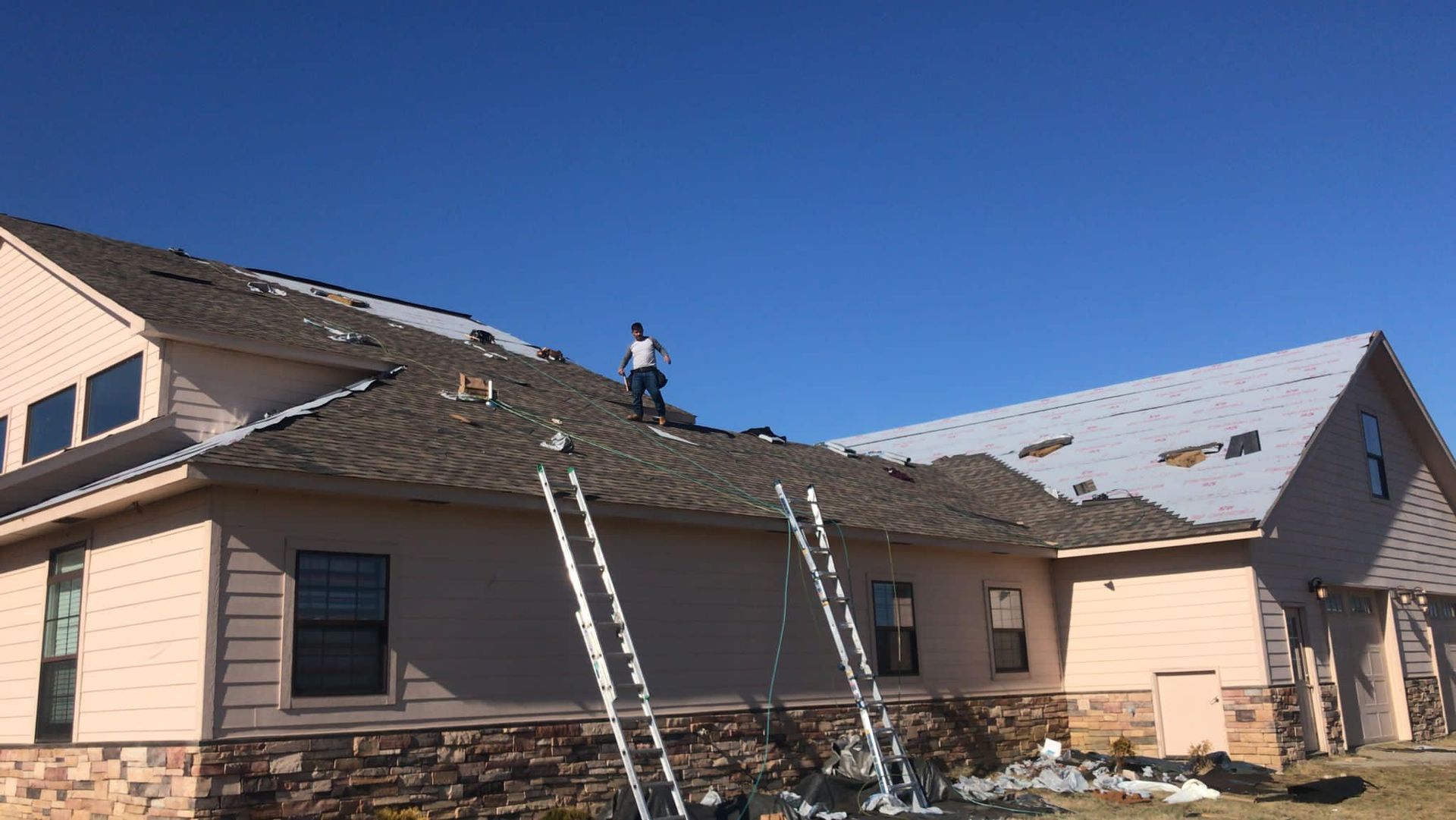 Workers on a house roof replacing shingles, with ladders leaning against the beige home under a clear blue sky