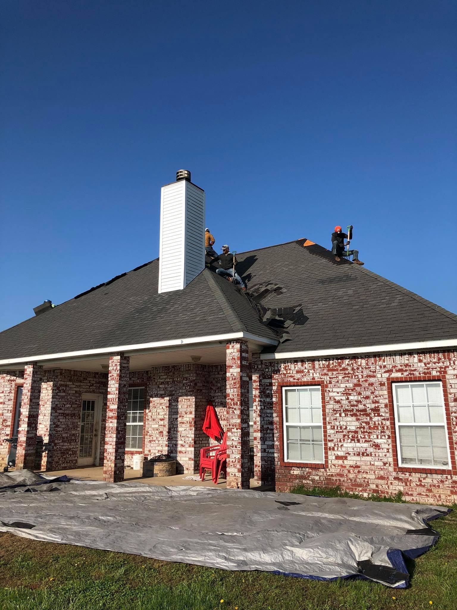 Brick house with a dark roof, white chimney, and construction materials on the roof under a clear blue sky