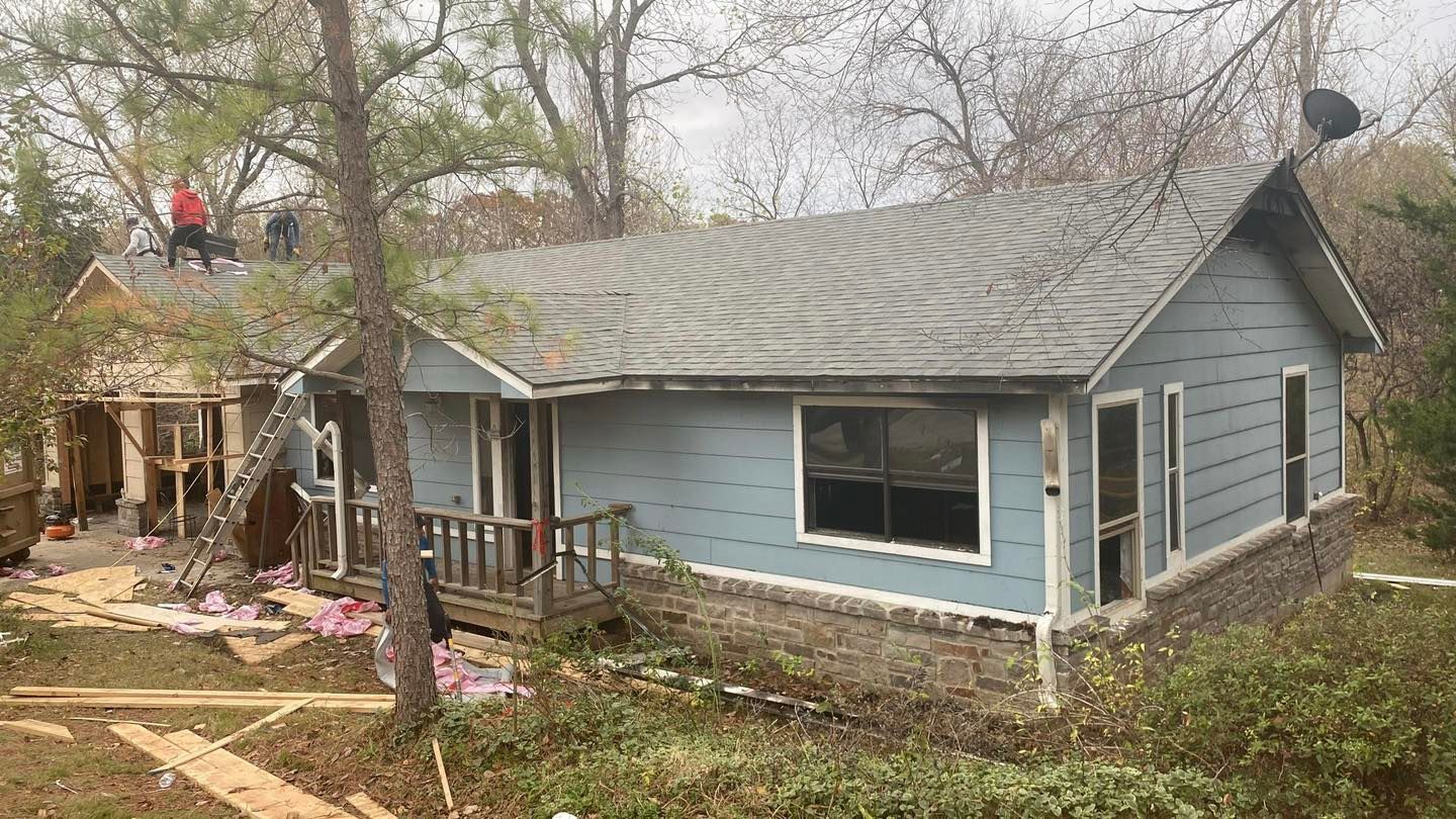 Damaged blue house with a collapsed porch and debris in a wooded yard