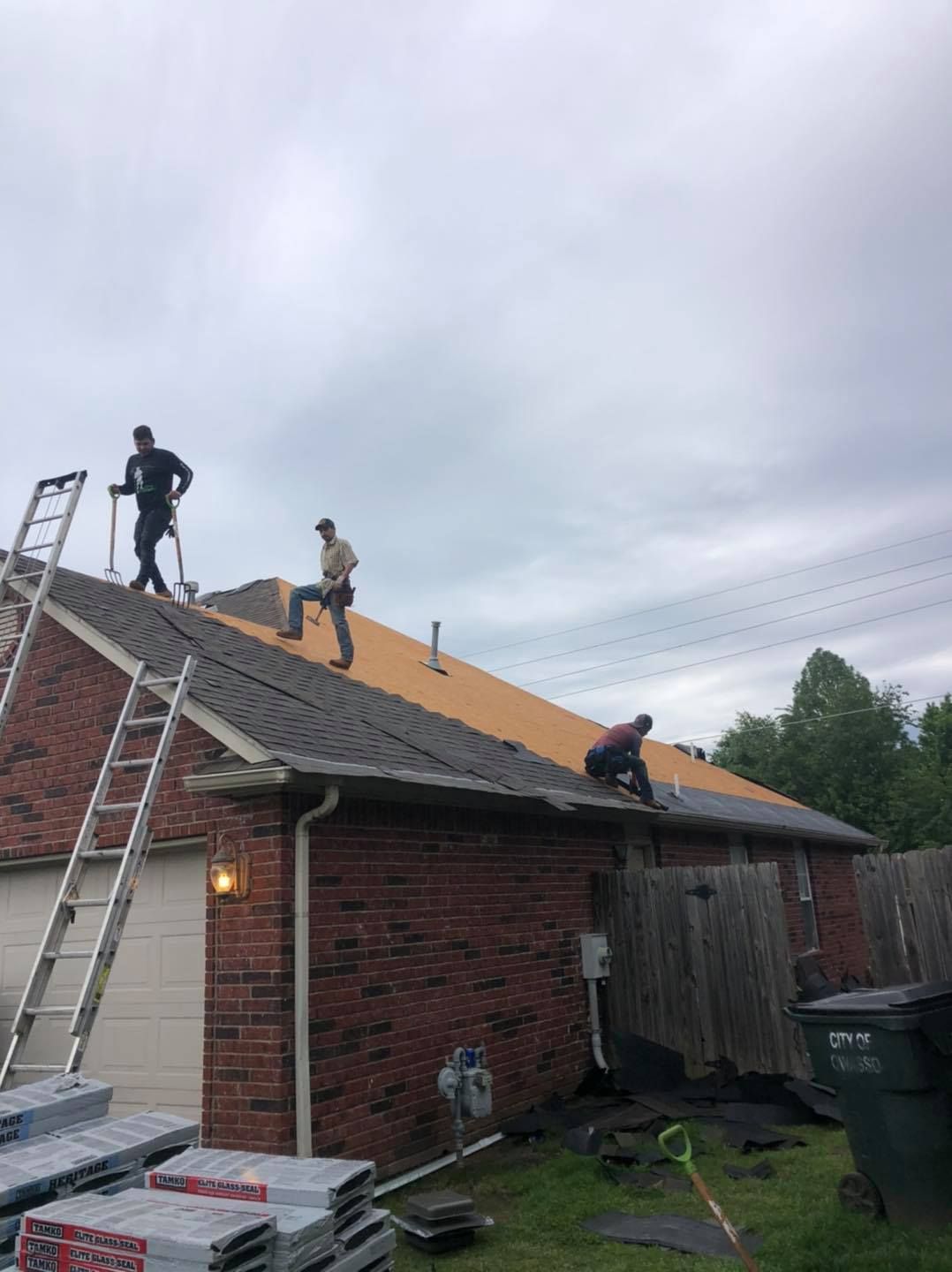 Workers roofing a brick house, with ladders and unfinished shingles under an overcast sky