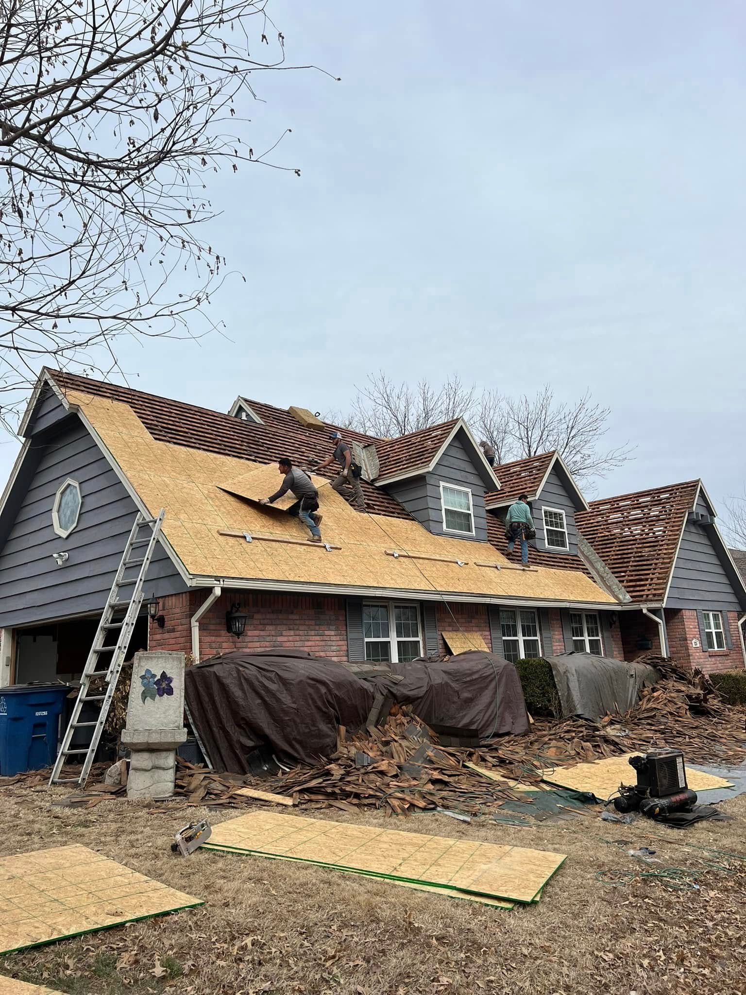 Storm-damaged house with torn roof shingles, debris in yard, and covered items along the side of the home