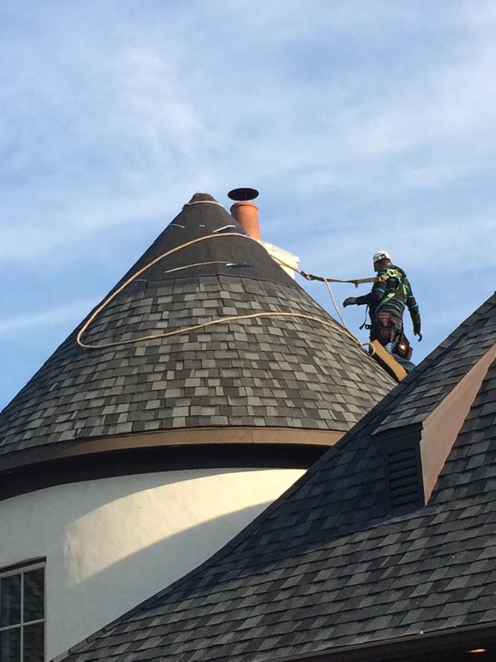Worker on a roof repairing dark shingle roof beside a round turret under a blue sky