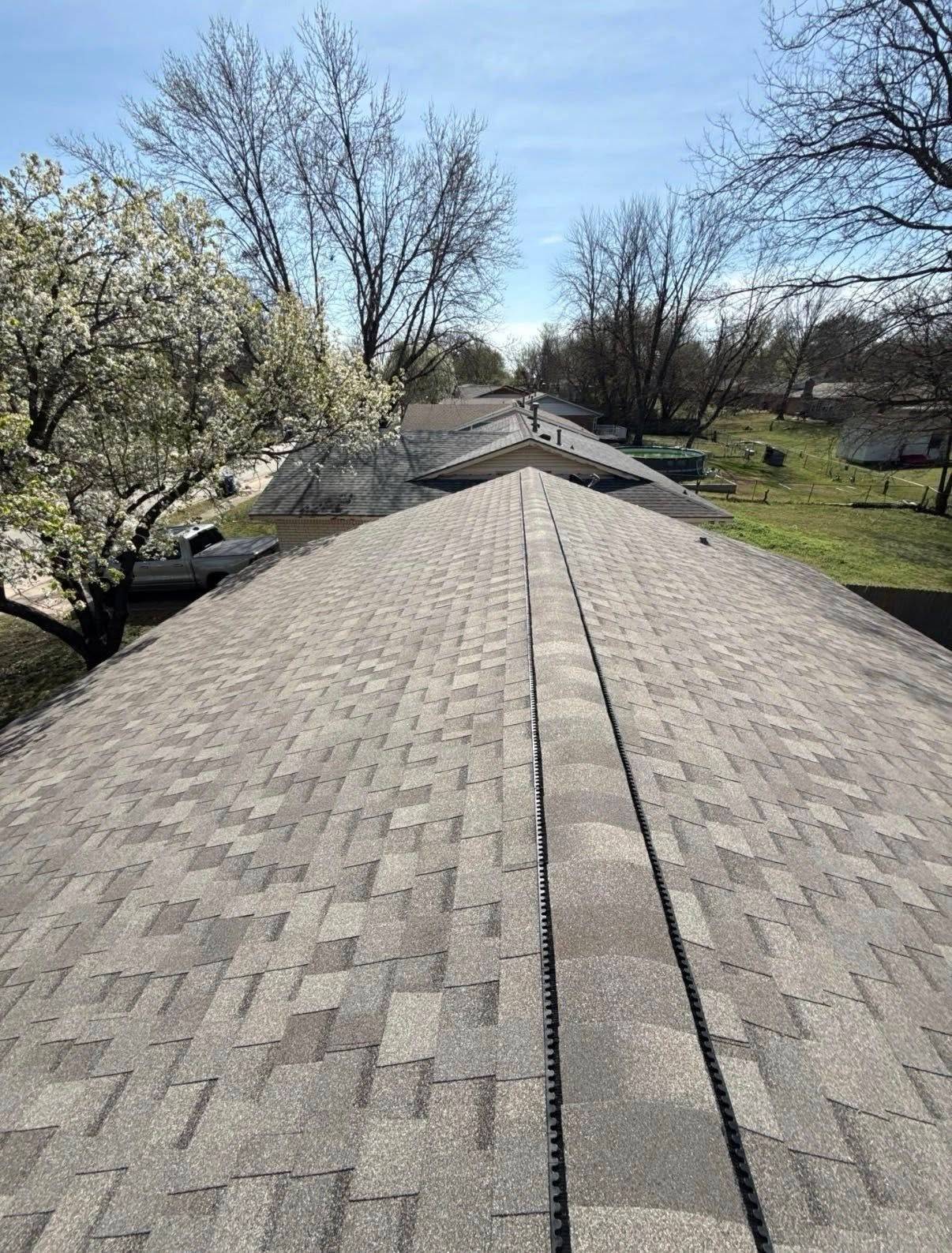Rooftop with gray shingles under a clear blue sky, bordered by bare trees and a grassy yard