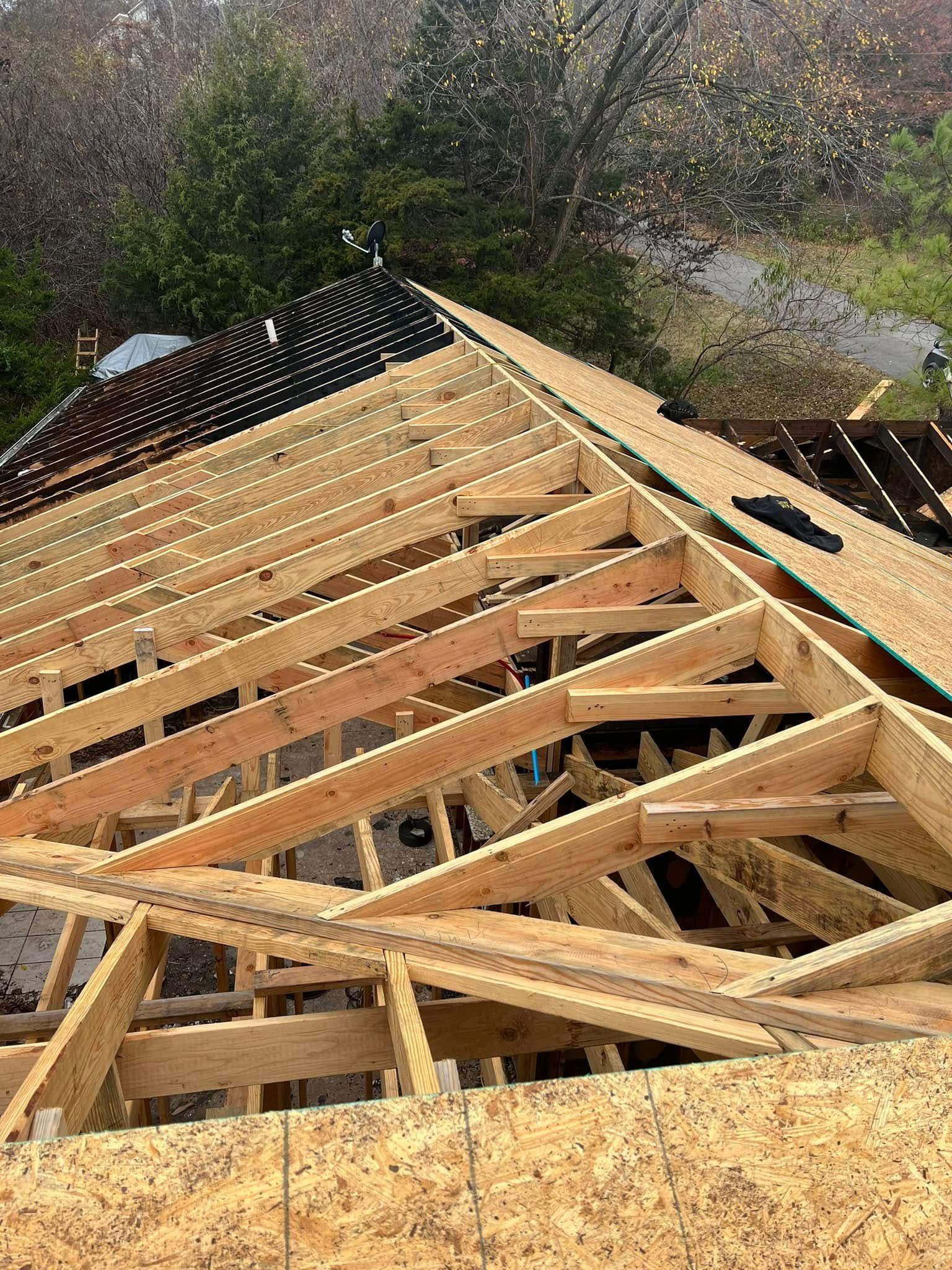 Wooden roof trusses under construction on a house, viewed from above amid trees and a wet roof.
