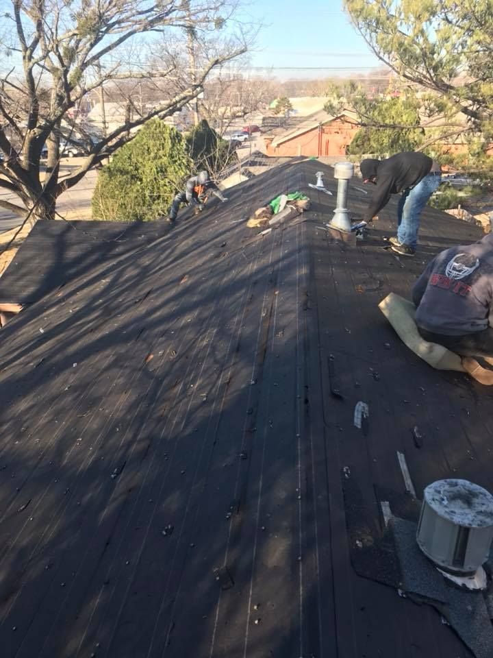 Workers repairing a dark roof on a sunny day, with trees and houses in the background