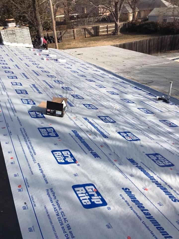 Roofer installing white underlayment on a sloped roof, with trees and a wooded hillside in the background